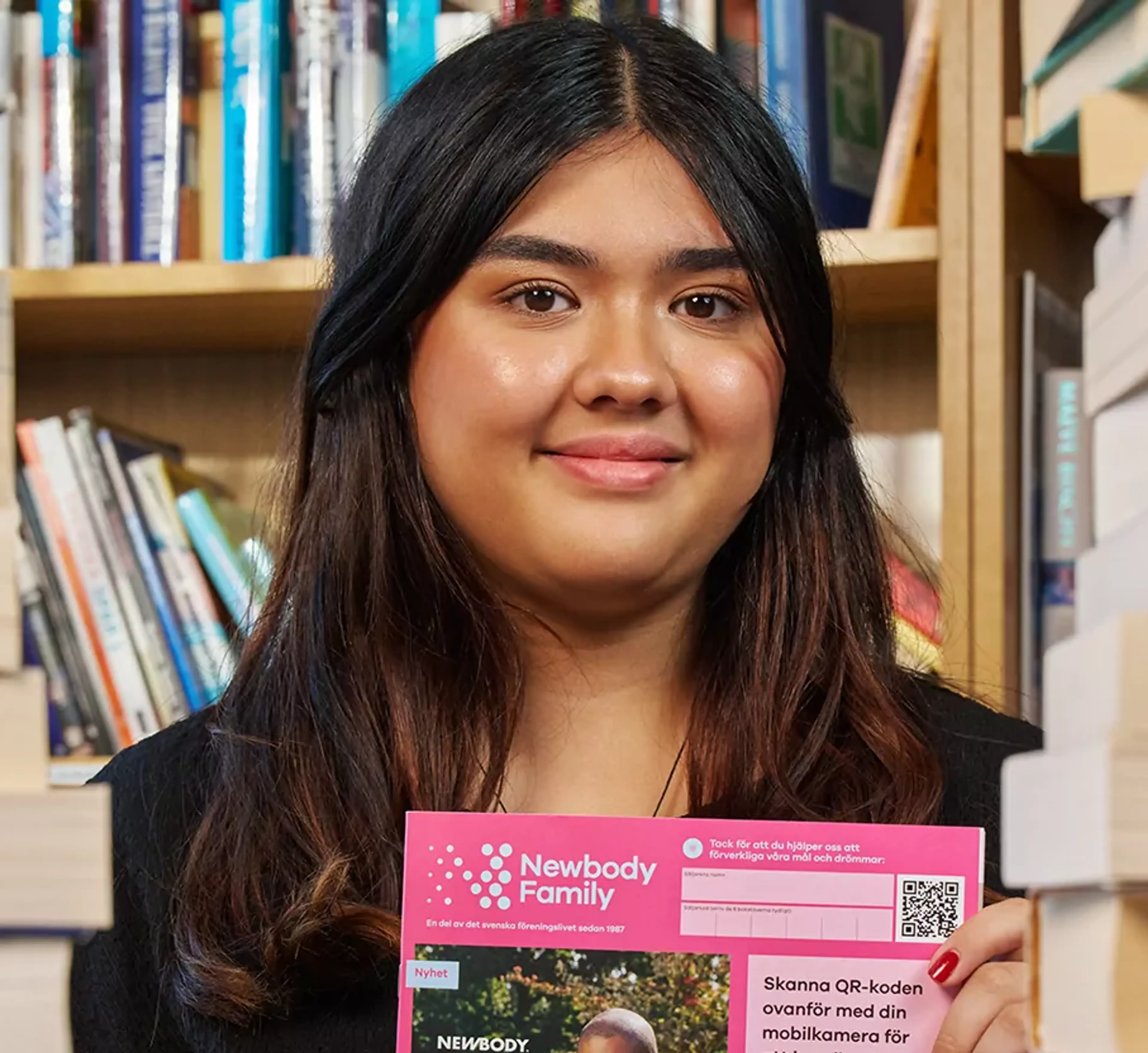 En ung tjej sitter mellan två högar med böcker och håller i en katalog med en rosa framsida från Newbody Family. I bakgrunden syns bokhyllor fyllda med böcker.