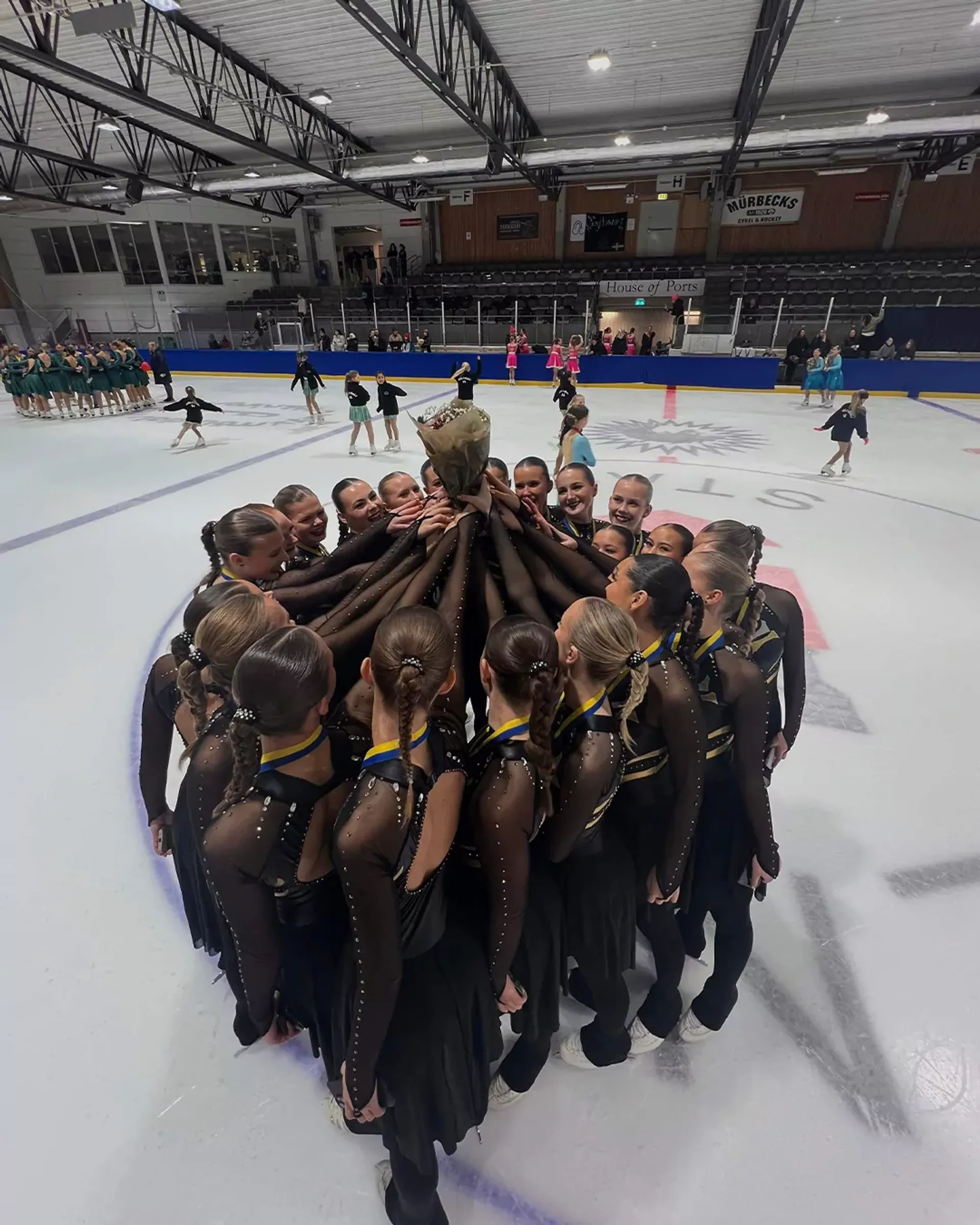 Ett konståkningsteam i svarta dräkter står tätt tillsammans i en cirkel på isen och håller upp en bukett blommor. I bakgrunden syns flera andra lag och tomma läktarsektioner i arenan.