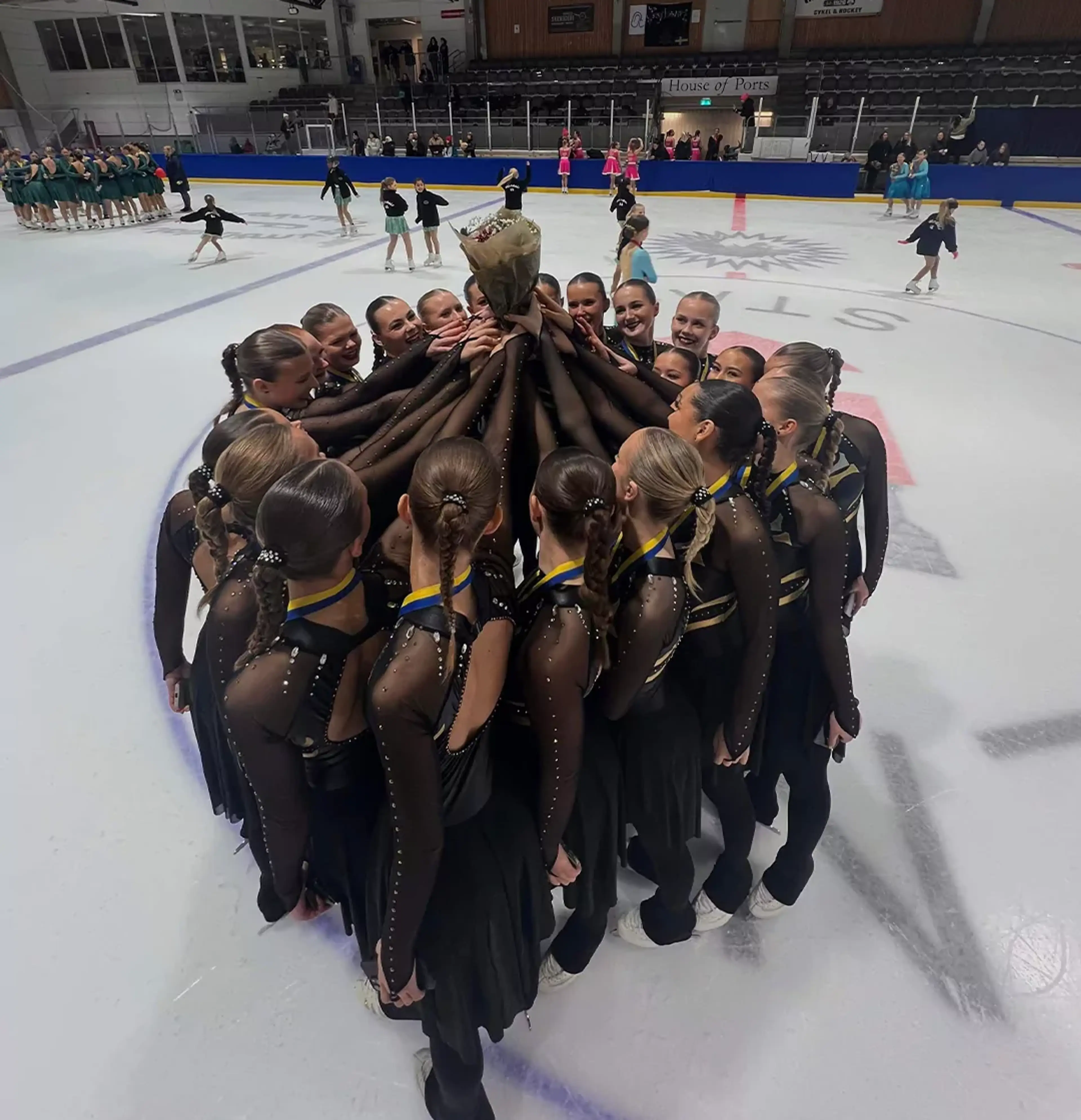 Ett konståkningsteam i svarta dräkter står tätt tillsammans i en cirkel på isen och håller upp en bukett blommor. I bakgrunden syns flera andra lag och tomma läktarsektioner i arenan.