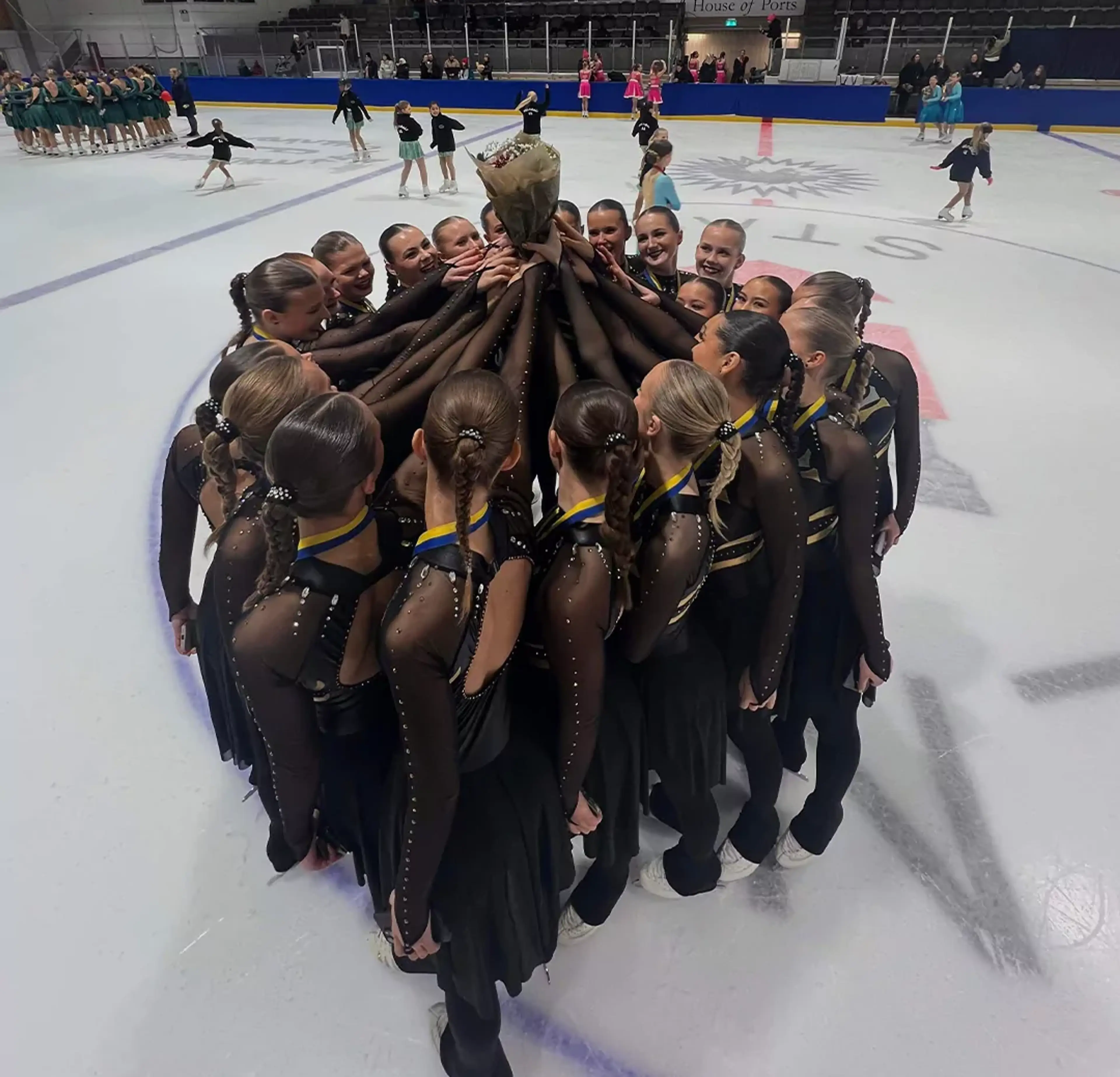 Ett konståkningsteam i svarta dräkter står tätt tillsammans i en cirkel på isen och håller upp en bukett blommor. I bakgrunden syns flera andra lag och tomma läktarsektioner i arenan.