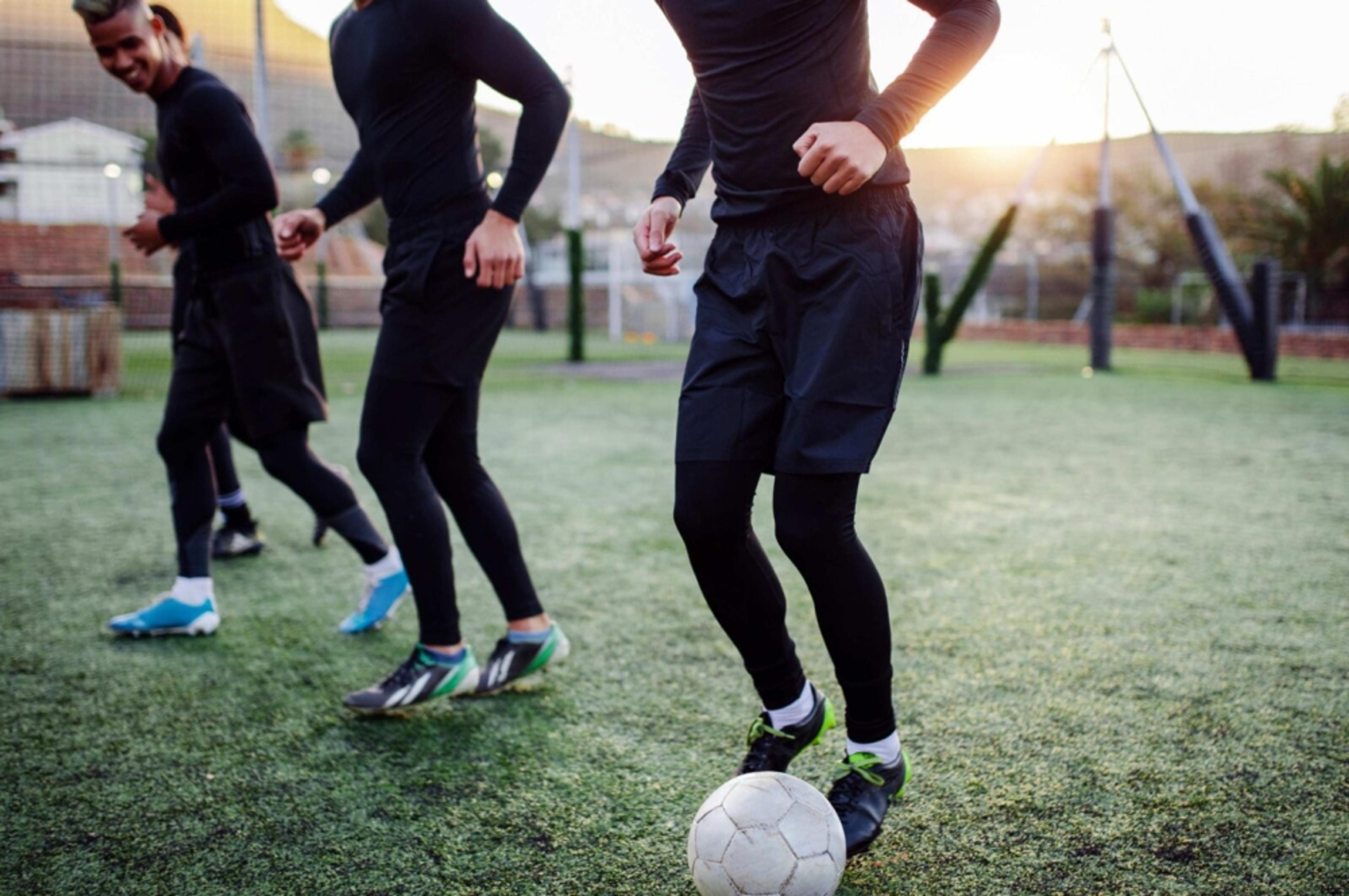 Fyra personer i sportkläder tränar på en gräsplan med en vit fotboll. Solen skiner bakom dem och skapar ett varmt ljus.