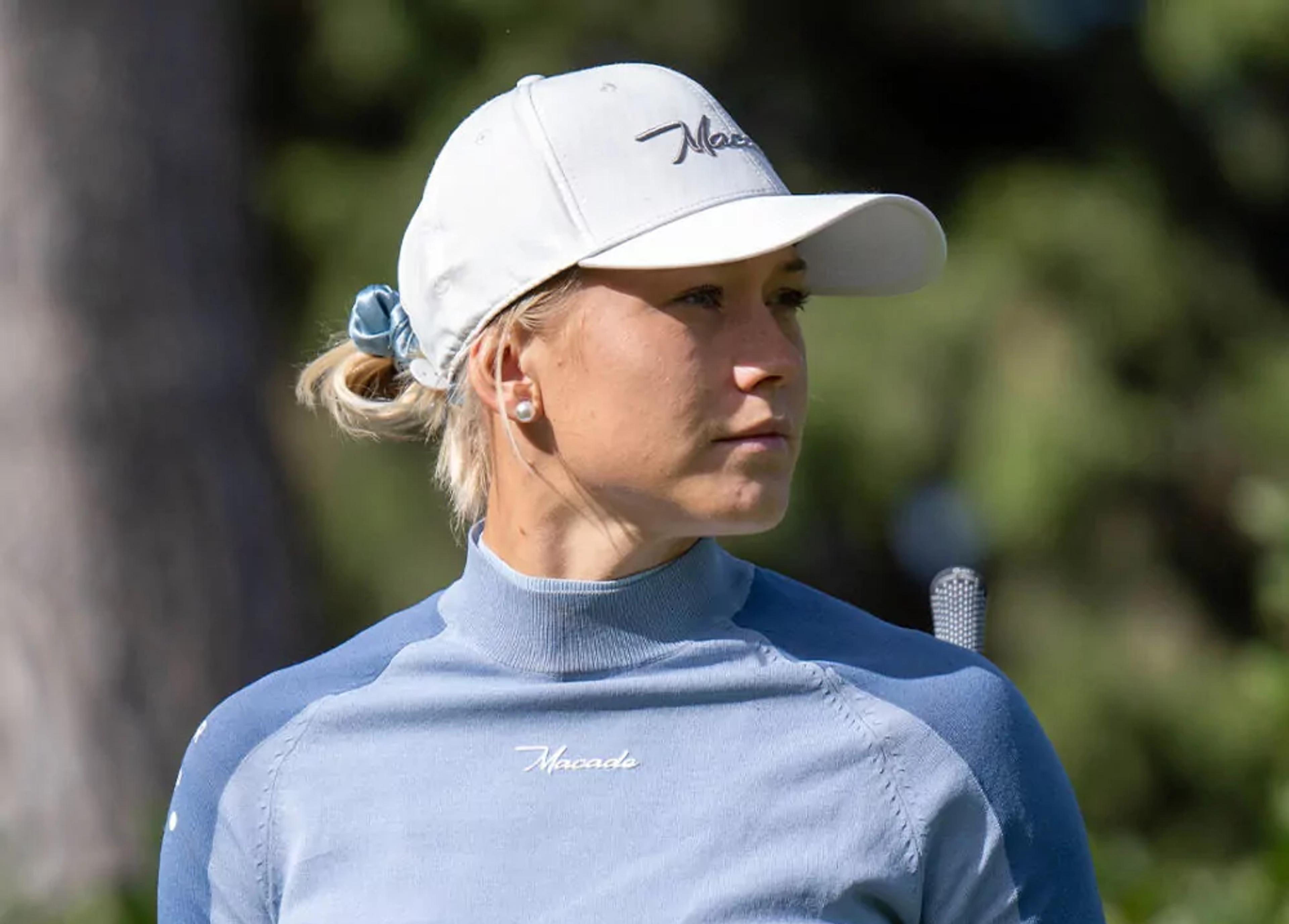 En kvinna i ljusblå golftröja och vit keps håller en golfklubba över axeln, med en lugn naturbakgrund.