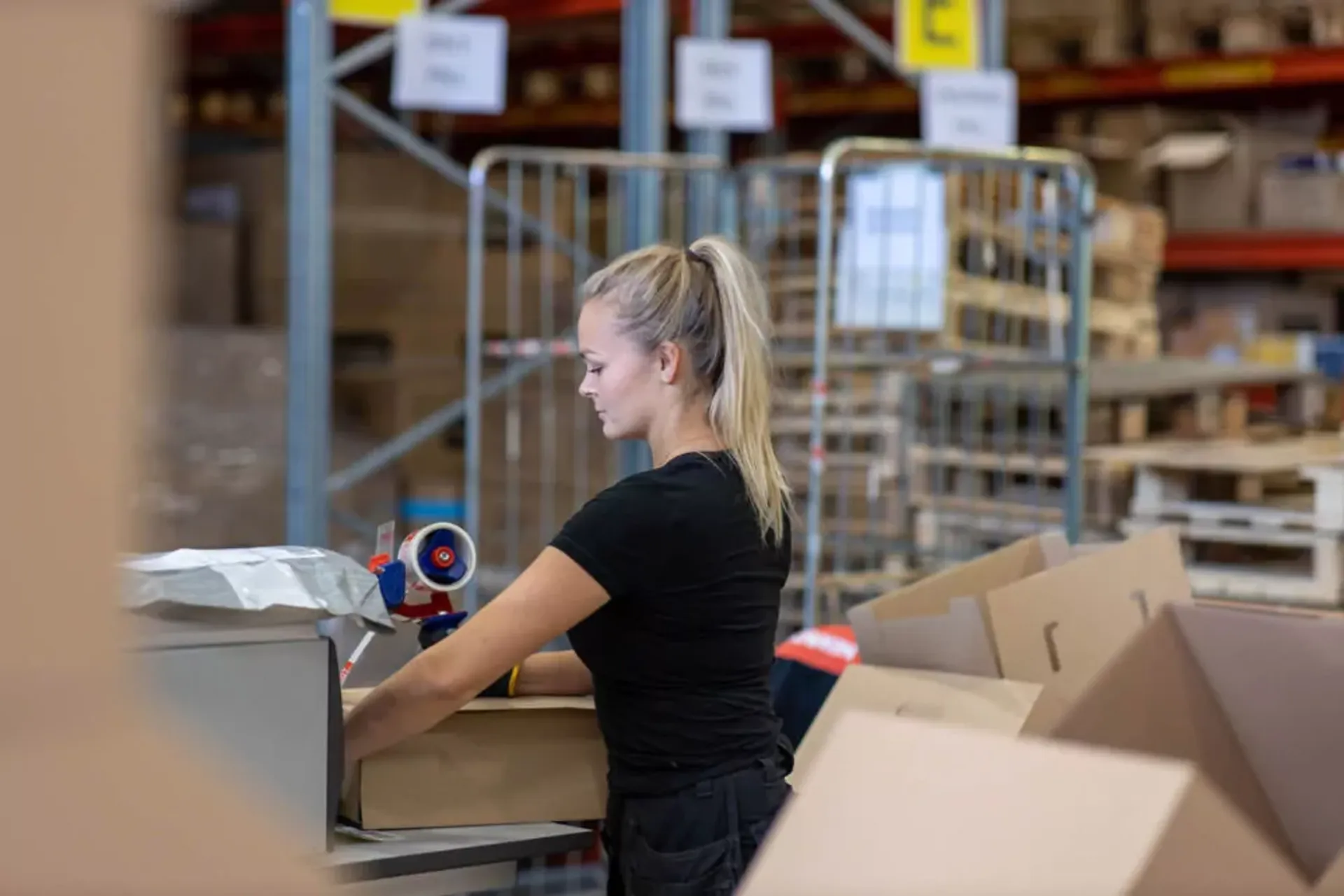 Kvinna som packar en kartong med hjälp av packtejp i ett lager fullt av paket och kartonger. Omkring henne syns hyllor och rullburar med fler varor.