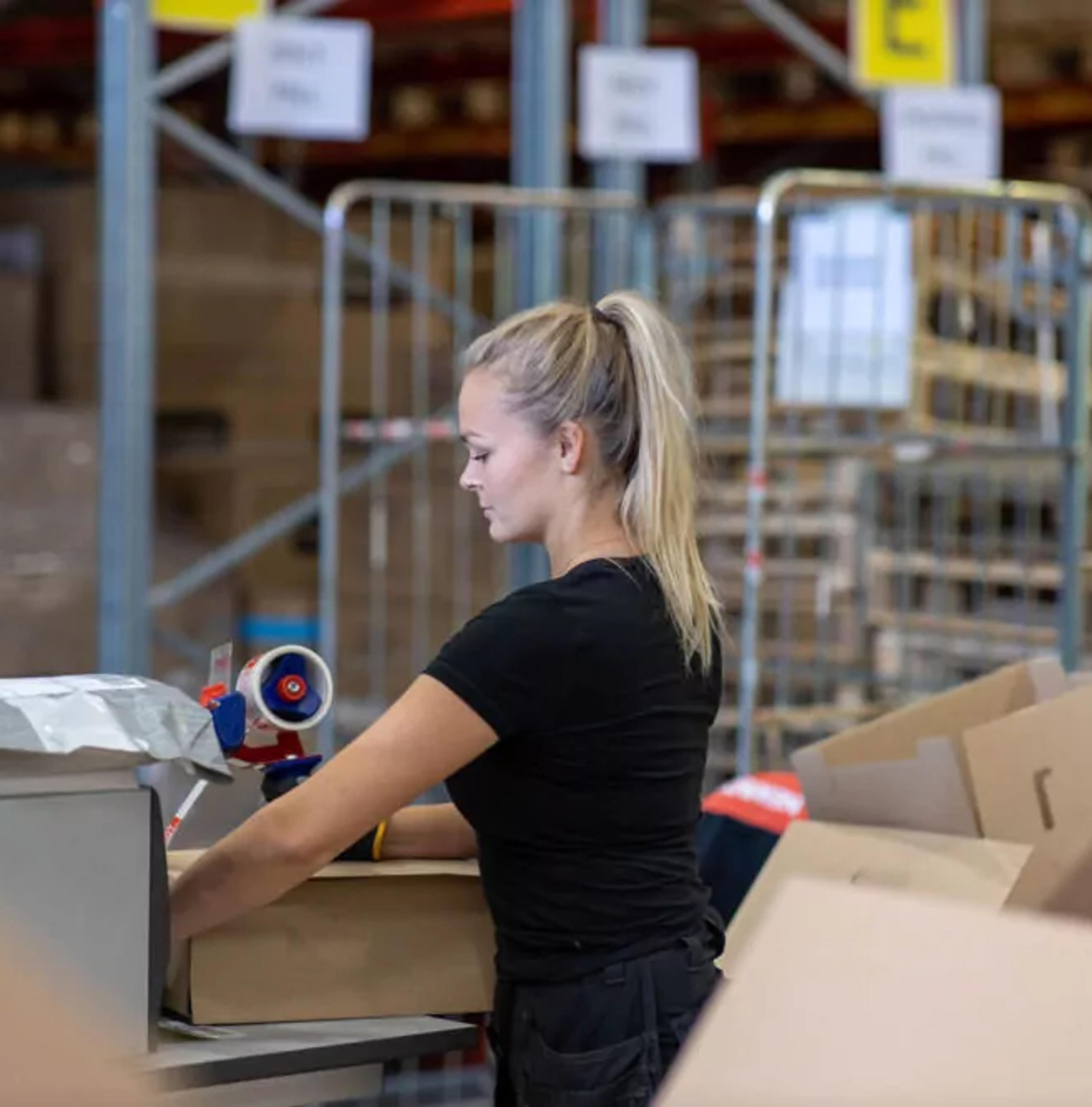 Kvinna som packar en kartong med hjälp av packtejp i ett lager fullt av paket och kartonger. Omkring henne syns hyllor och rullburar med fler varor.