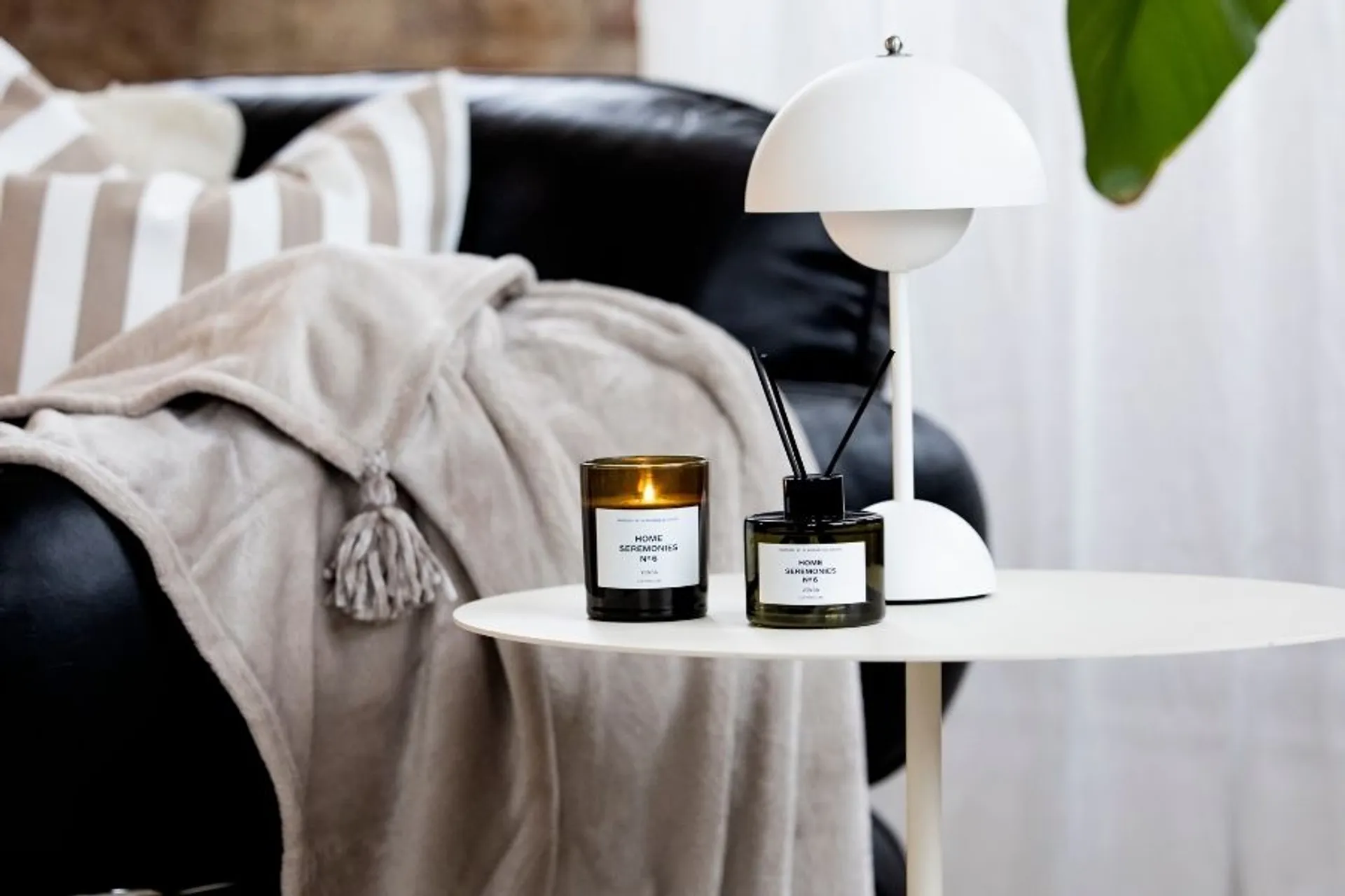 Doftljuset och rumsdoften i Aroma Set Vinda på sidobord med vit lampa bredvid svart skinnsoffa med filt