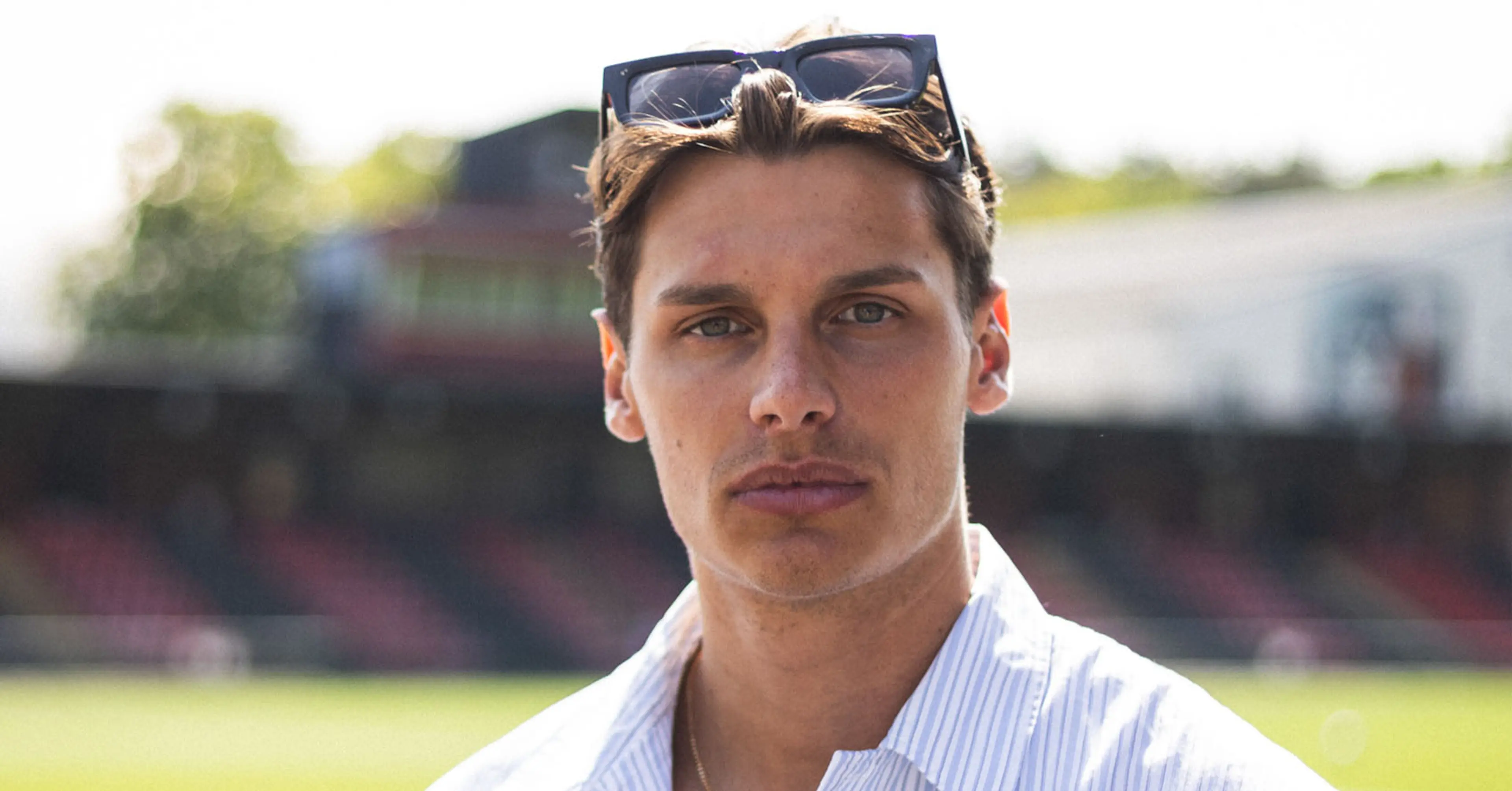 En man i ljusblå skjorta och solglasögon i håret står vid ett stängsel med en fotbollsplan i bakgrunden. Ljuset är mjukt och det är en solig dag.