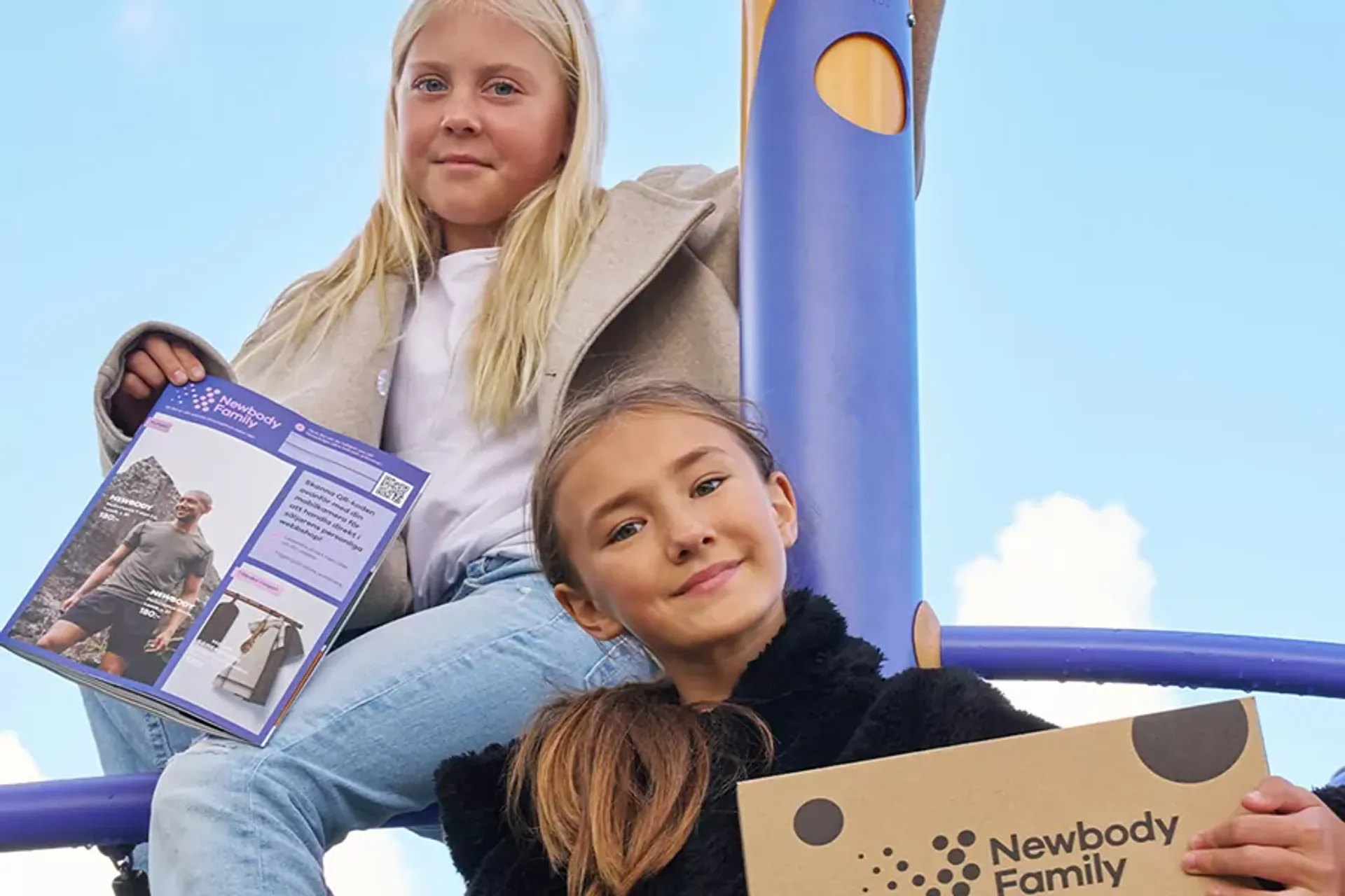 Två flickor sitter på en lekställning med en katalog och en kartong i händerna. De ser glada och trygga ut under en klarblå himmel.