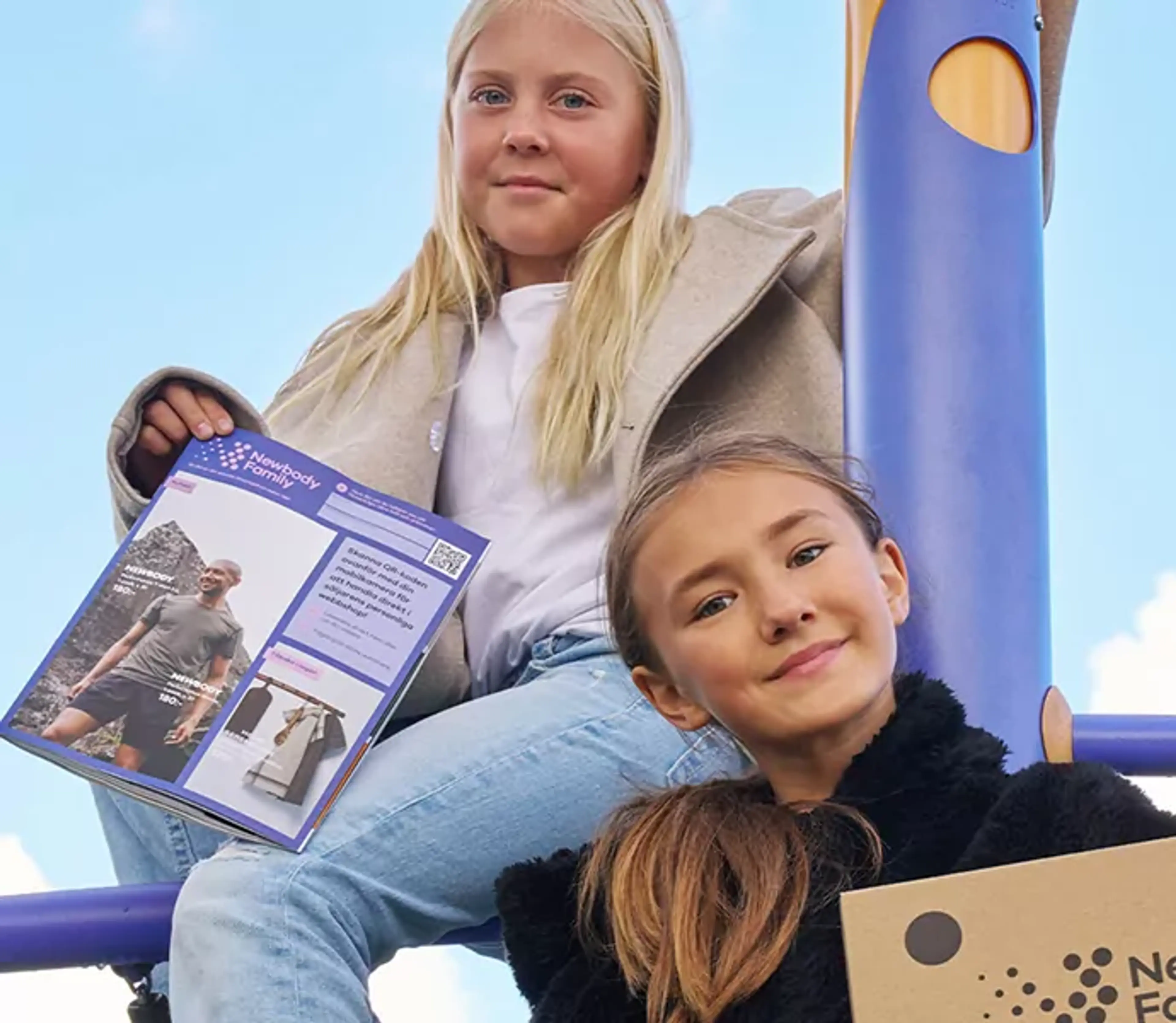Två flickor sitter på en lekställning med en katalog och en kartong i händerna. De ser glada och trygga ut under en klarblå himmel.