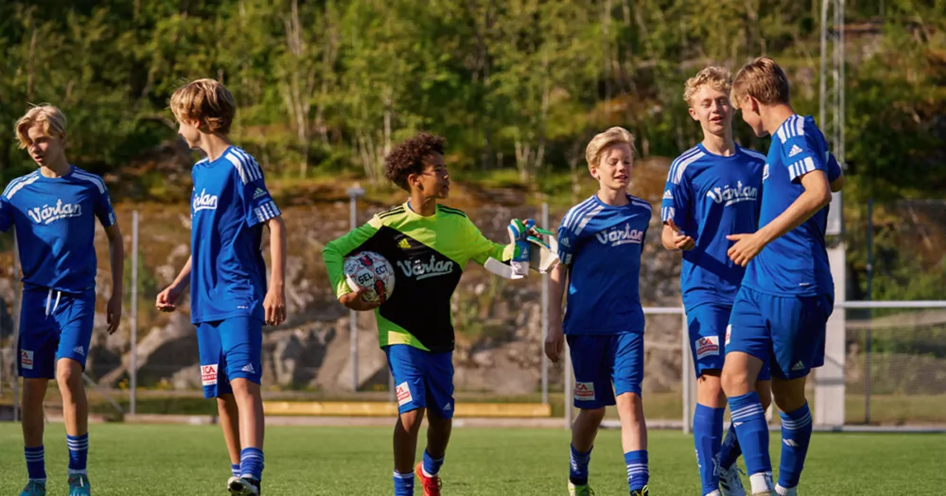 En grupp ungdomsspelare i blå fotbollsdräkter på en utomhusplan, med en målvakt i grön och svart tröja som håller en fotboll. Bakgrunden visar grönska och stenpartier.
