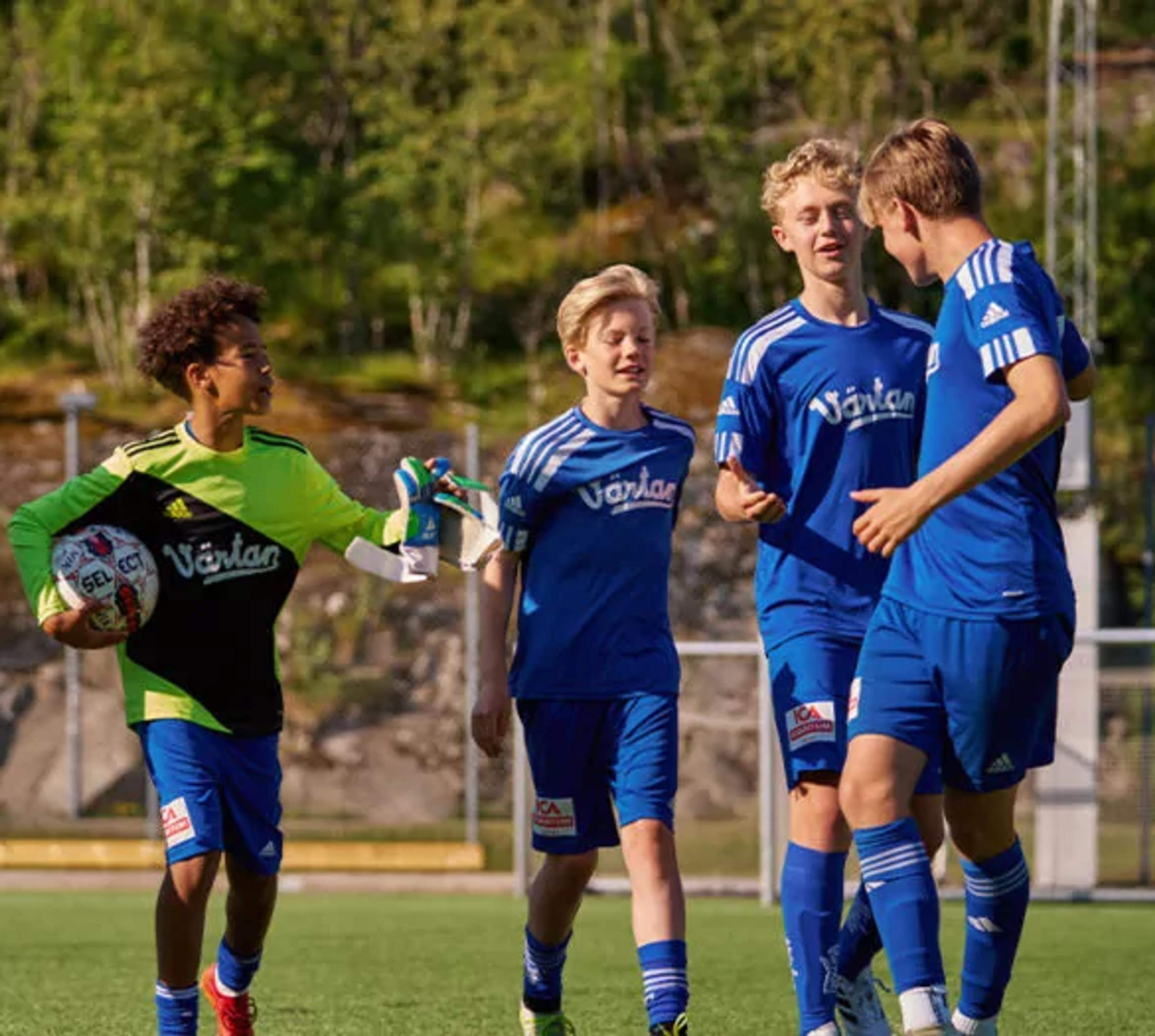 En grupp ungdomsspelare i blå fotbollsdräkter på en utomhusplan, med en målvakt i grön och svart tröja som håller en fotboll. Bakgrunden visar grönska och stenpartier.
