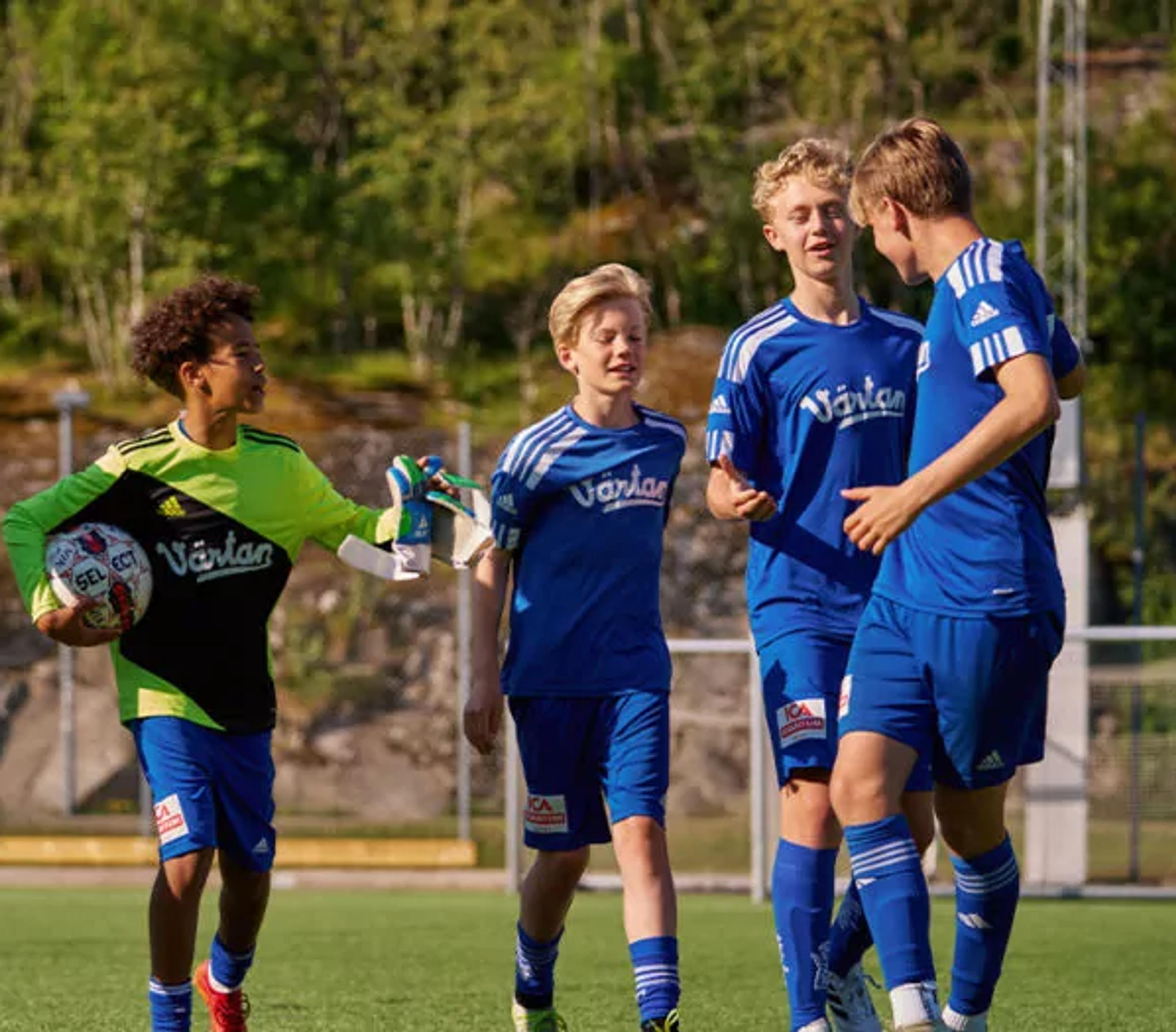 En grupp ungdomsspelare i blå fotbollsdräkter på en utomhusplan, med en målvakt i grön och svart tröja som håller en fotboll. Bakgrunden visar grönska och stenpartier.