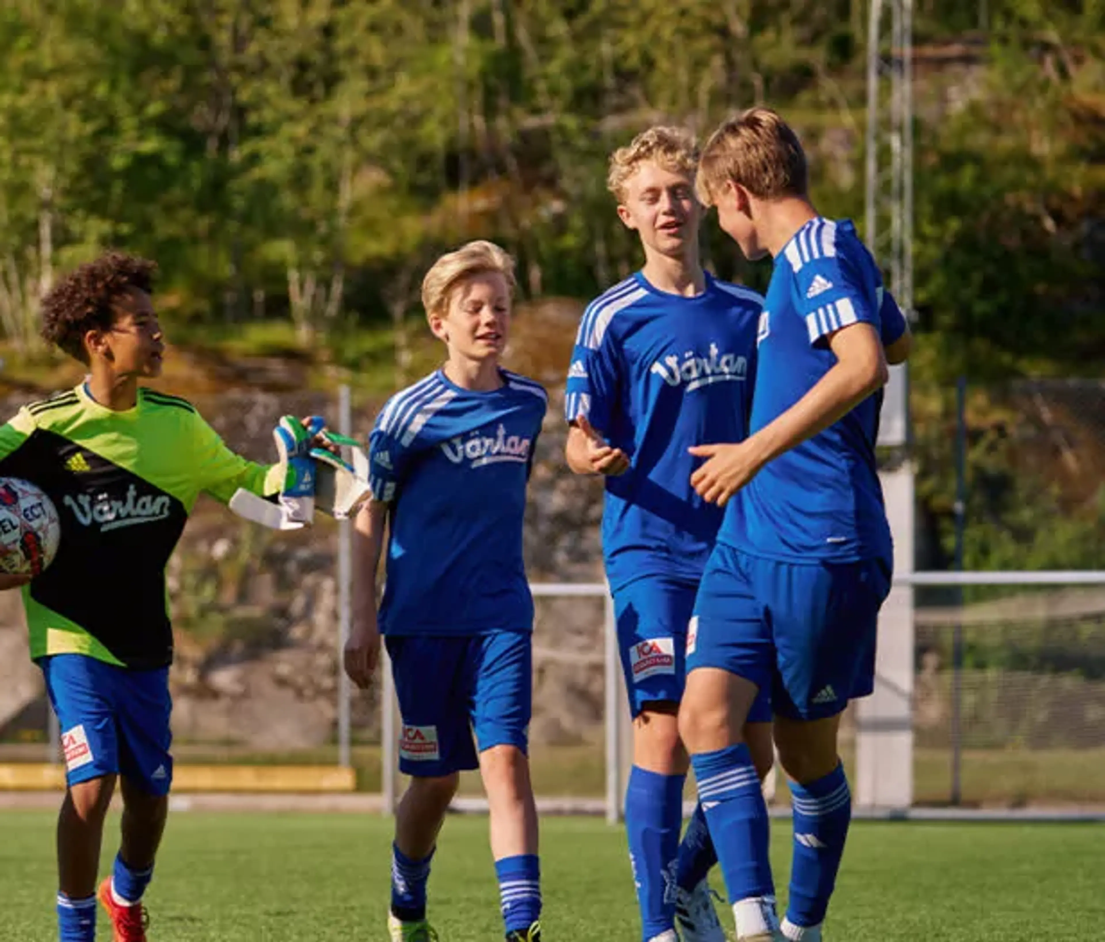 En grupp ungdomsspelare i blå fotbollsdräkter på en utomhusplan, med en målvakt i grön och svart tröja som håller en fotboll. Bakgrunden visar grönska och stenpartier.