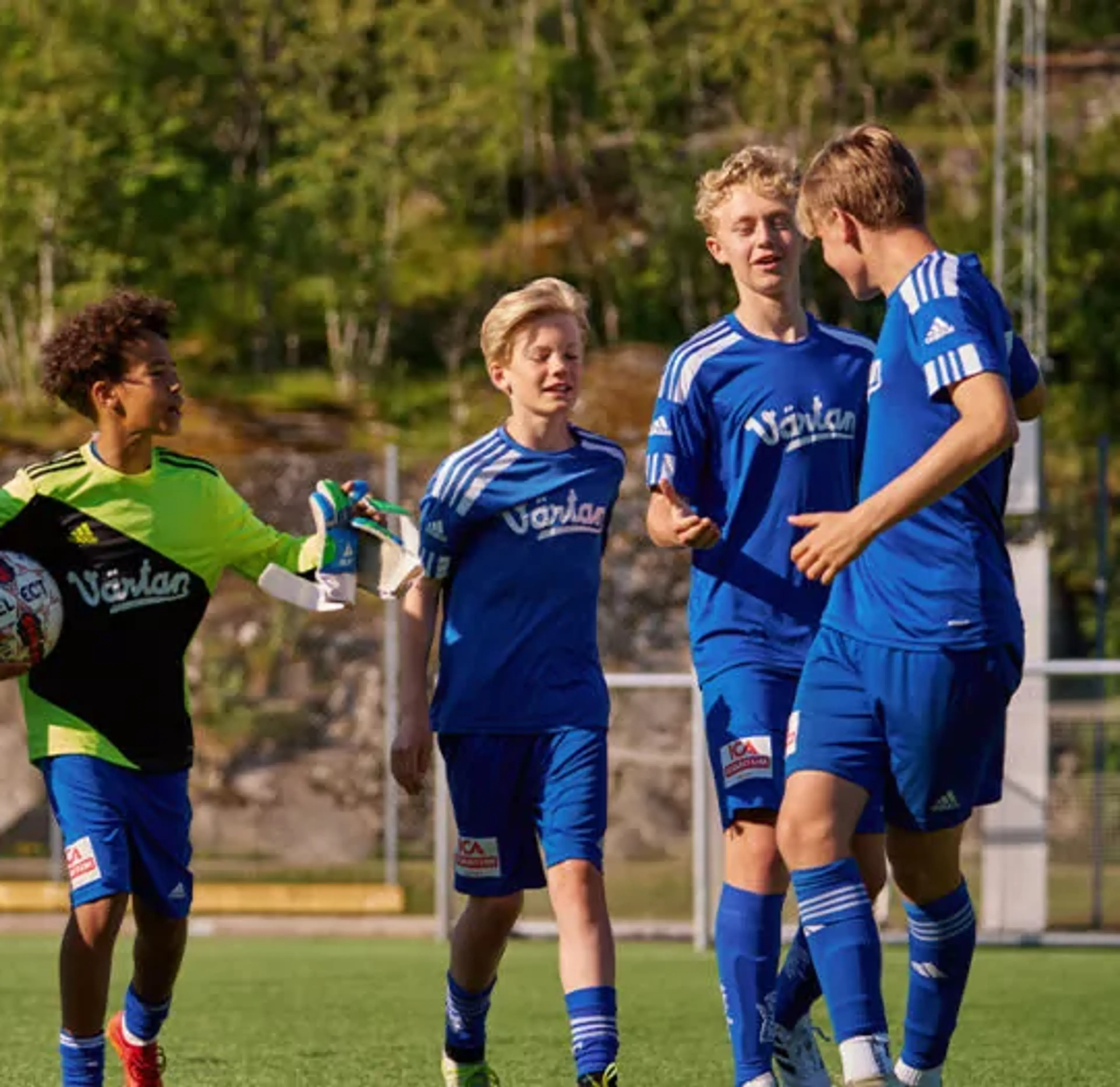 En grupp ungdomsspelare i blå fotbollsdräkter på en utomhusplan, med en målvakt i grön och svart tröja som håller en fotboll. Bakgrunden visar grönska och stenpartier.