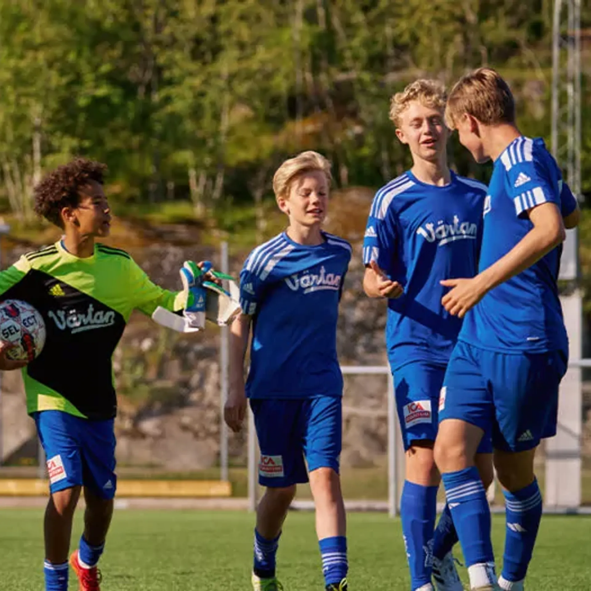 En grupp ungdomsspelare i blå fotbollsdräkter på en utomhusplan, med en målvakt i grön och svart tröja som håller en fotboll. Bakgrunden visar grönska och stenpartier.