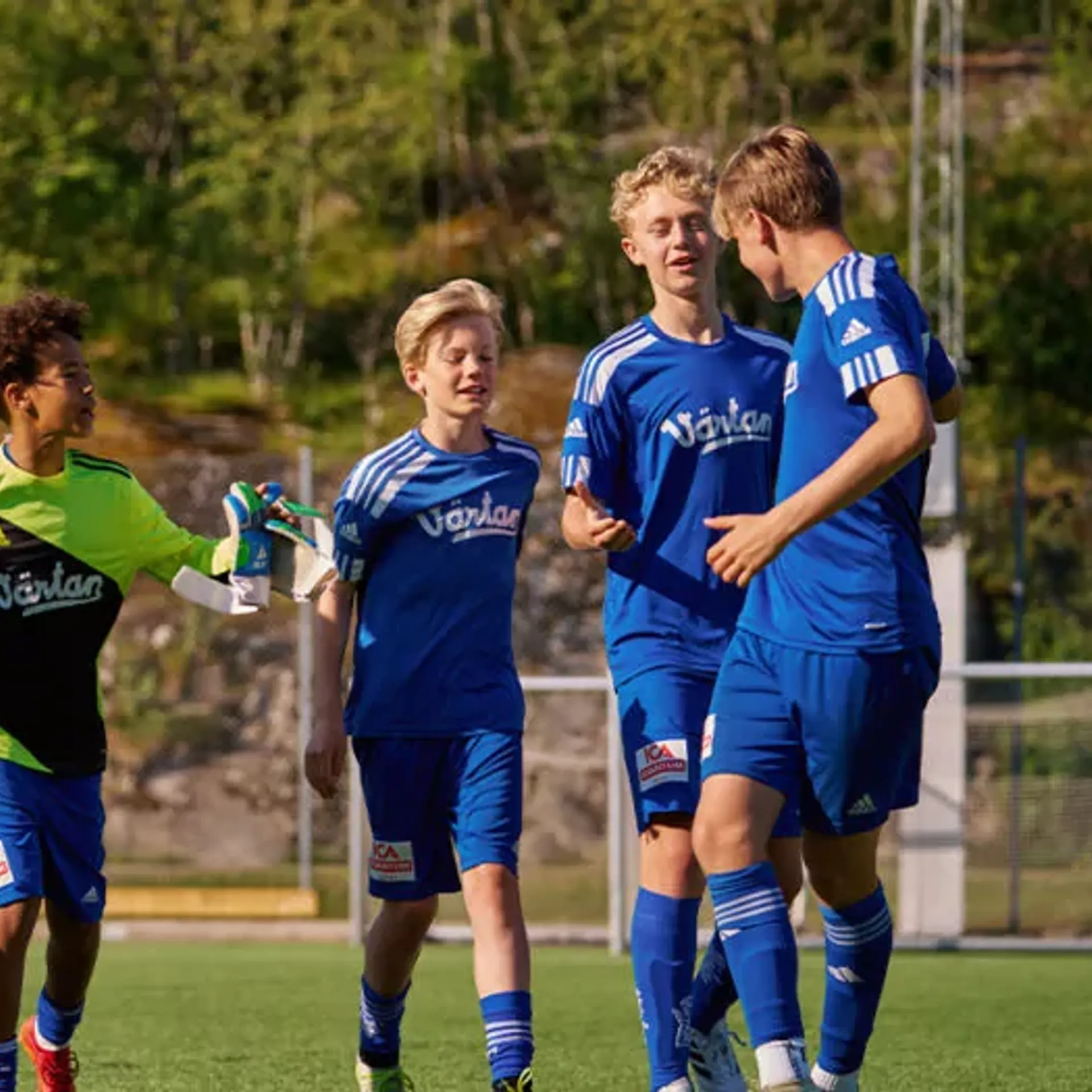 En grupp ungdomsspelare i blå fotbollsdräkter på en utomhusplan, med en målvakt i grön och svart tröja som håller en fotboll. Bakgrunden visar grönska och stenpartier.