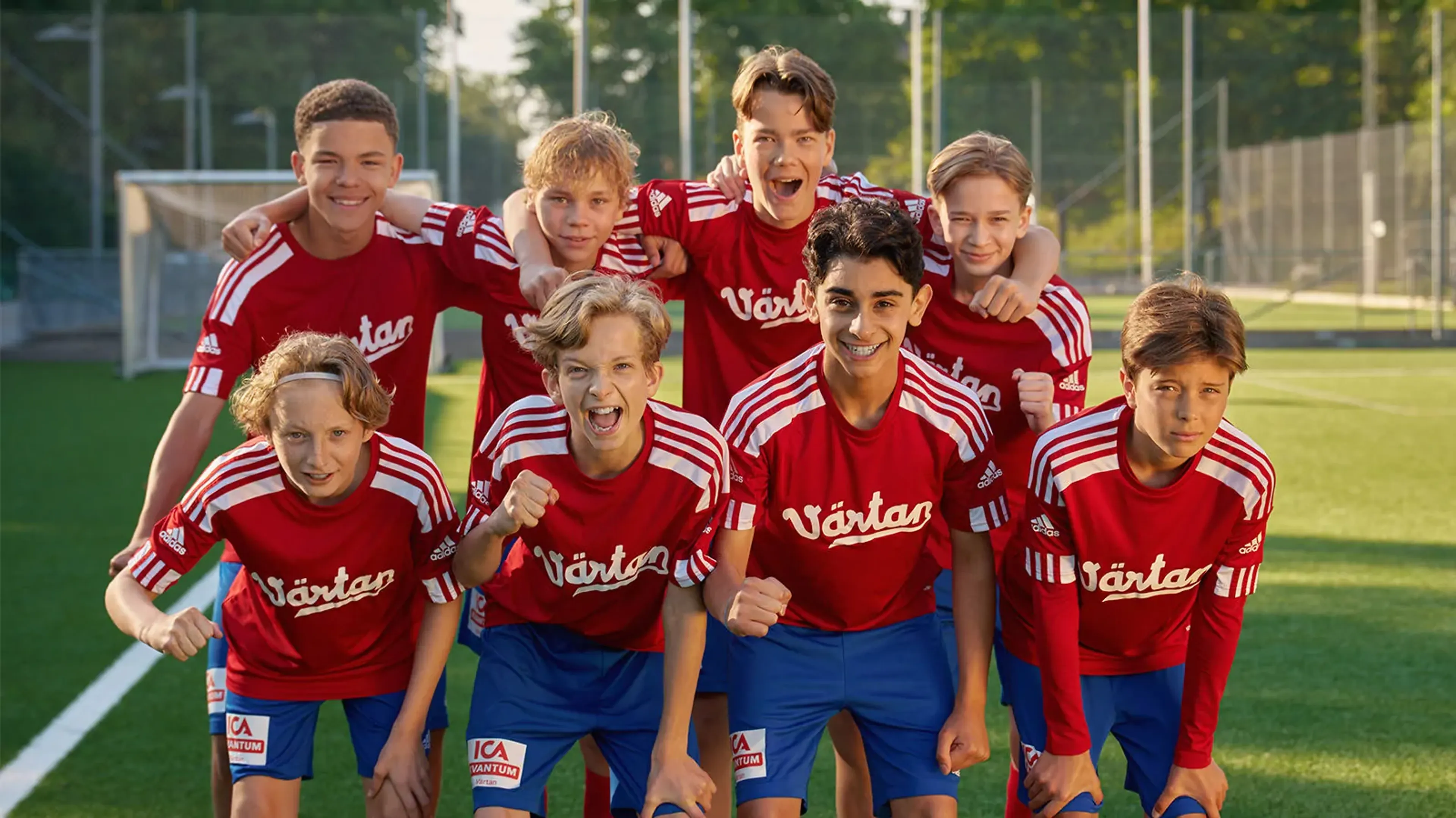 En grupp killar i röda tröjor och blå shorts står samlade på en fotbollsplan, vissa med armarna om varandra. De ser ivriga och taggade ut inför match eller träning.