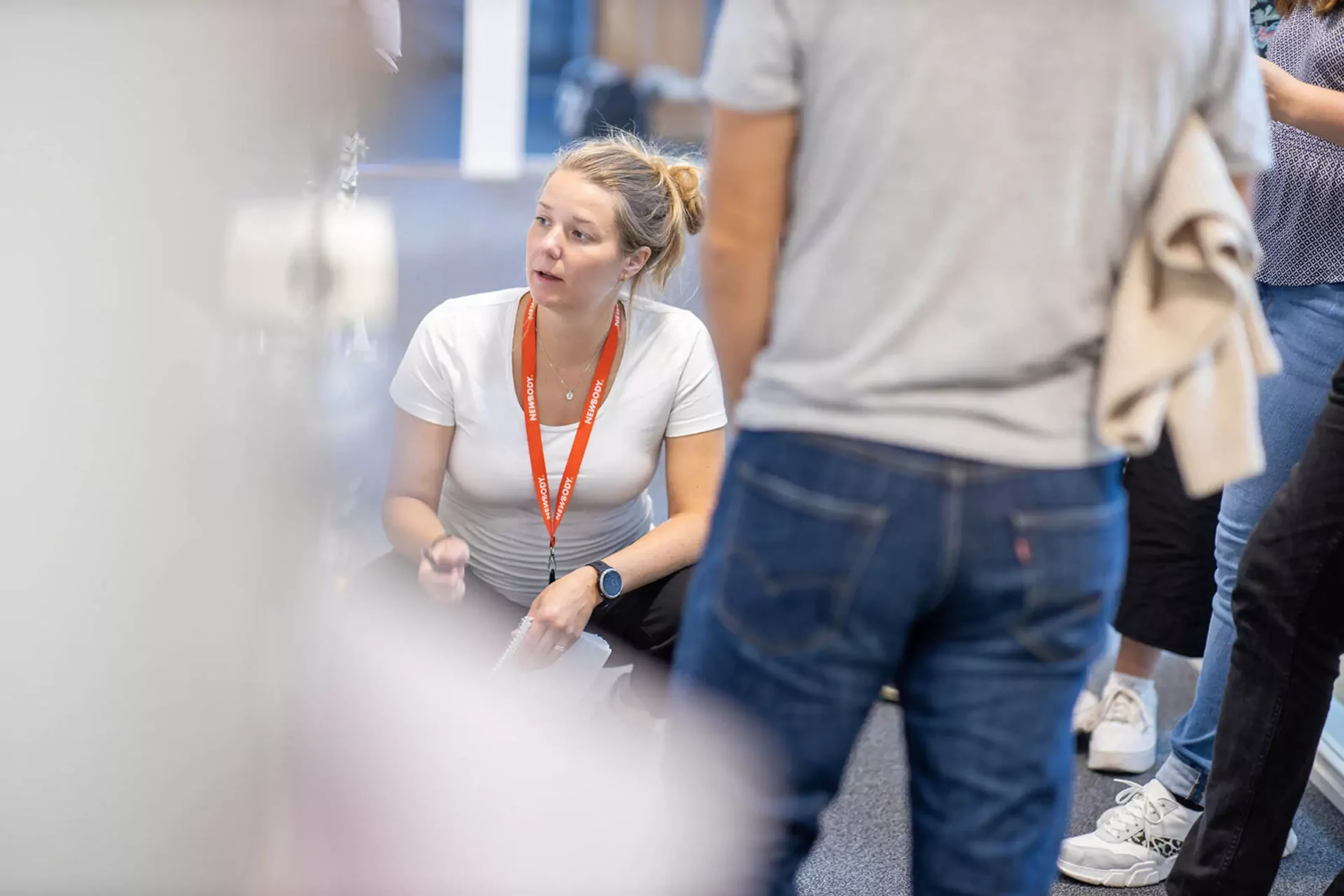 En kvinna i vit t-shirt och orange namnbricka sitter på huk och samtalar med flera personer som står runt omkring. Fokus är på mötet och den engagerade samtalsstilen.
