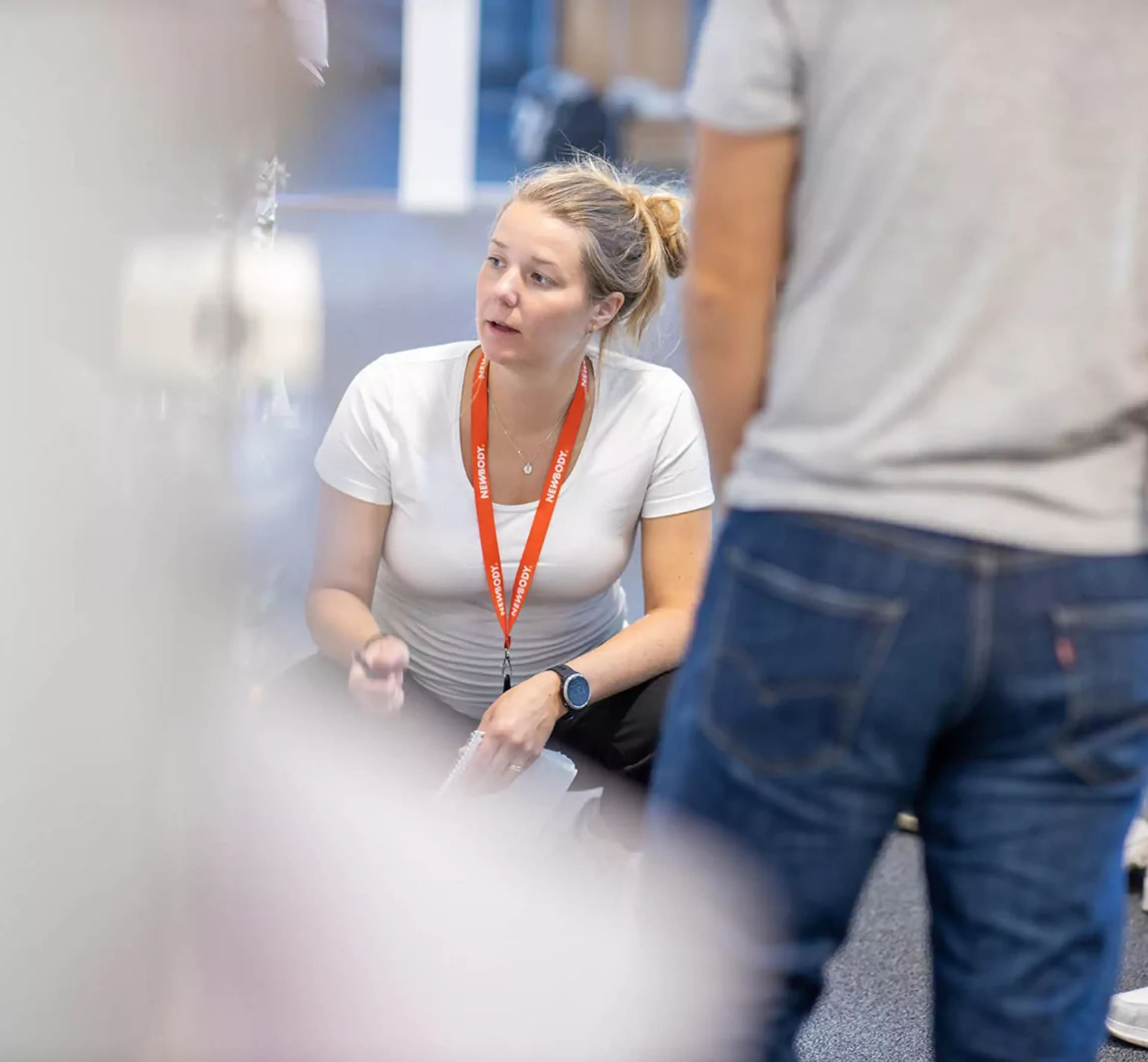 En kvinna i vit t-shirt och orange namnbricka sitter på huk och samtalar med flera personer som står runt omkring. Fokus är på mötet och den engagerade samtalsstilen.