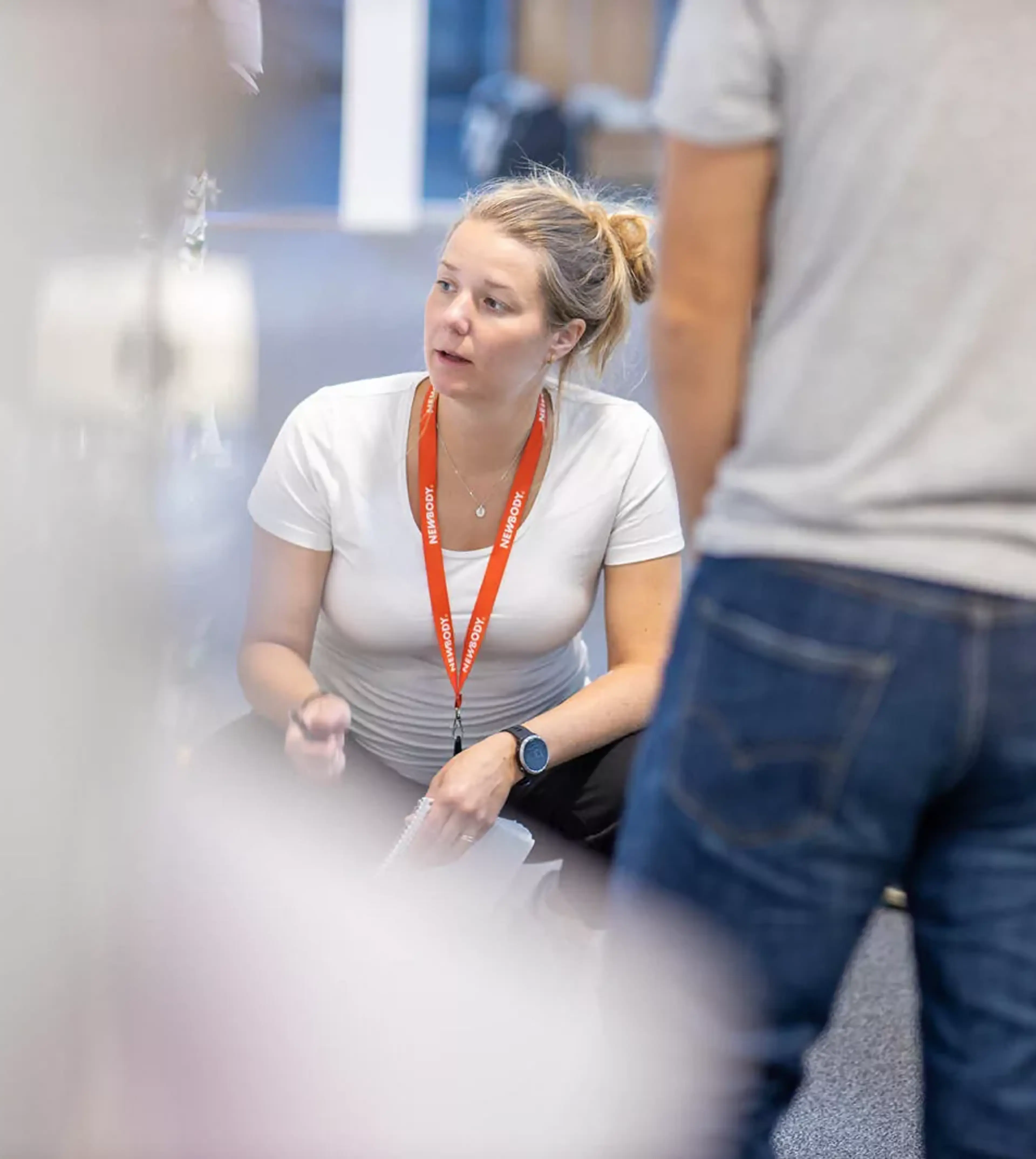 En kvinna i vit t-shirt och orange namnbricka sitter på huk och samtalar med flera personer som står runt omkring. Fokus är på mötet och den engagerade samtalsstilen.