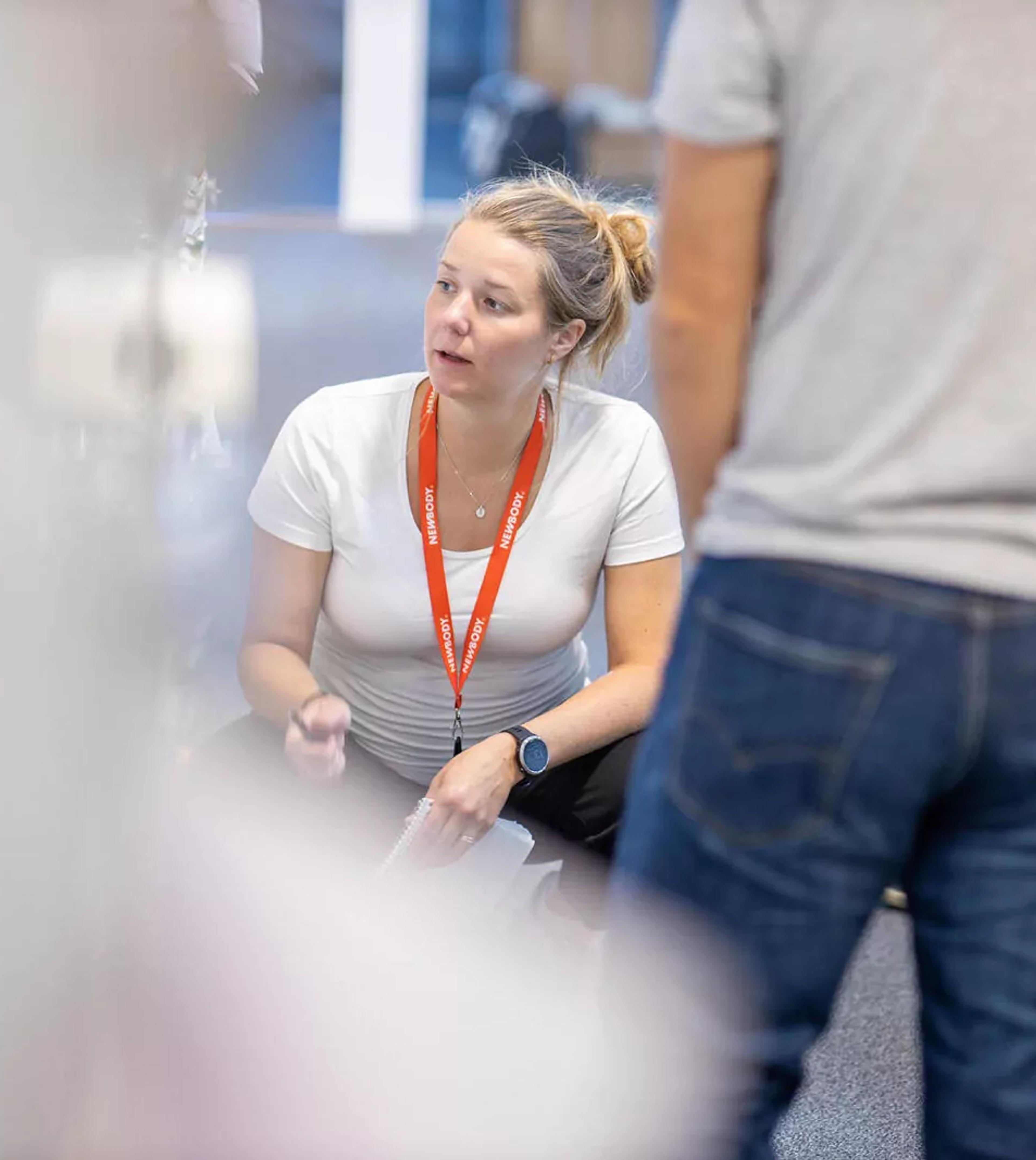 En kvinna i vit t-shirt och orange namnbricka sitter på huk och samtalar med flera personer som står runt omkring. Fokus är på mötet och den engagerade samtalsstilen.