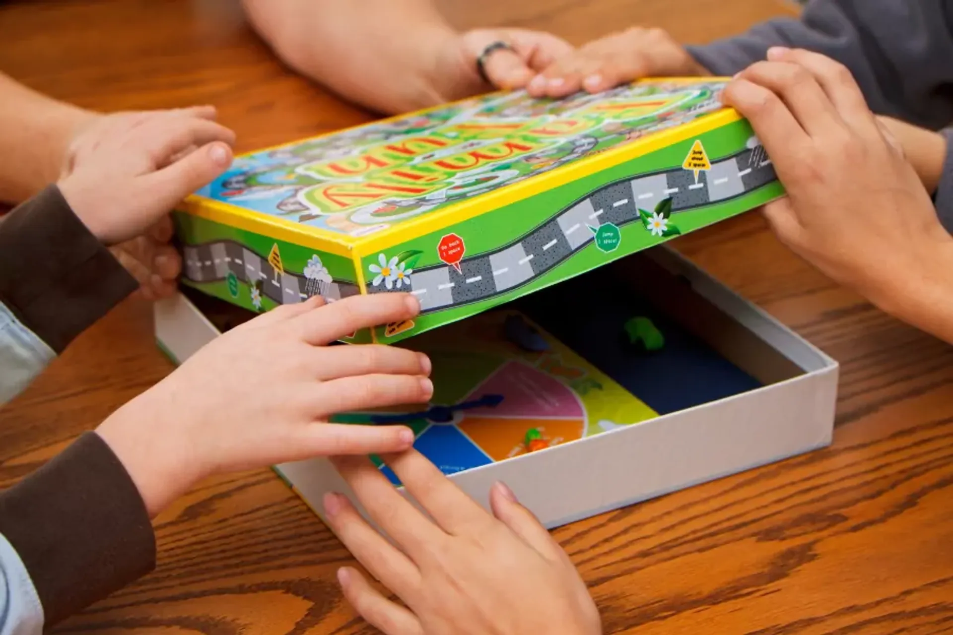 Flera händer håller på att öppna en färgglad spelask med ett brädspel inuti. Spelet har en vägillustration på locket och spelpjäser syns i botten av asken.