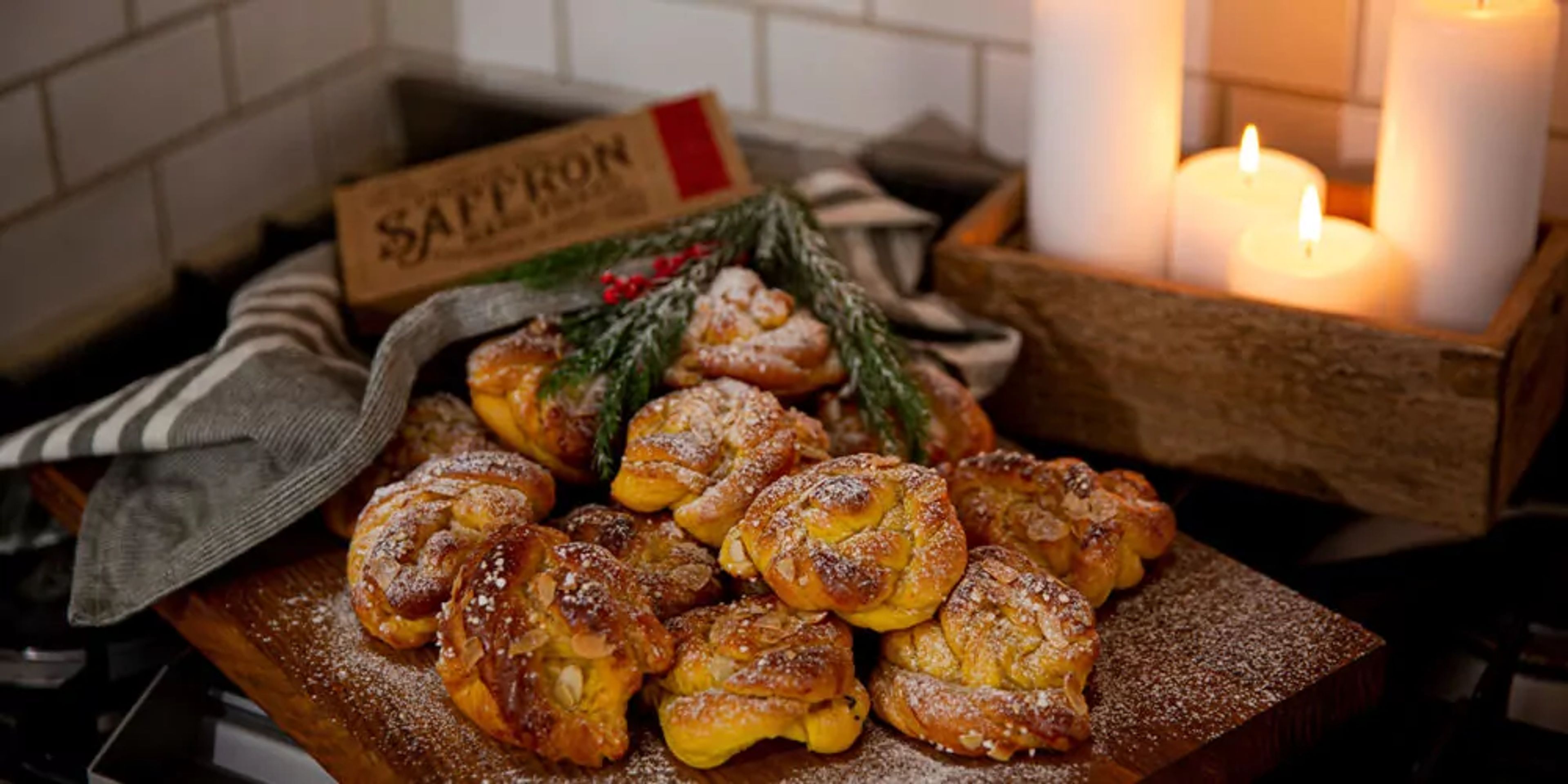 Ett fat med nybakade saffransbullar, pudrade med florsocker och dekorerade med mandelspån. I bakgrunden tända ljus och en saffranspåse.