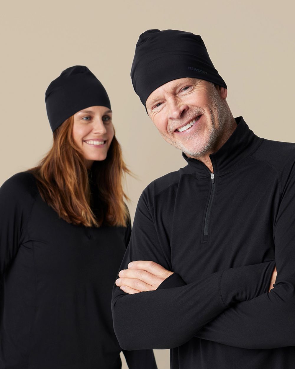 Två personer i svarta, enkla och släta mössor i lite elastiskt material. Båda bär svarta tröjor med hög krage, en med dragkedja.