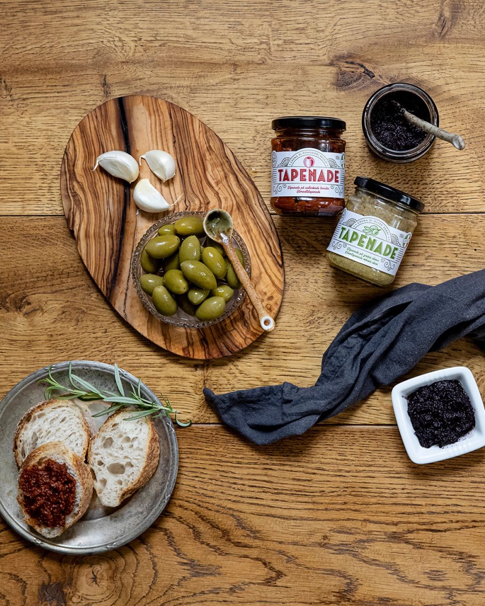 Två glasburkar med tapenade, en skål med gröna oliver och vitlöksklyftor på ett träfat. Ett fat med brödskivor, en burk och en liten skål med mörk tapenade samt en grå duk på ett trä