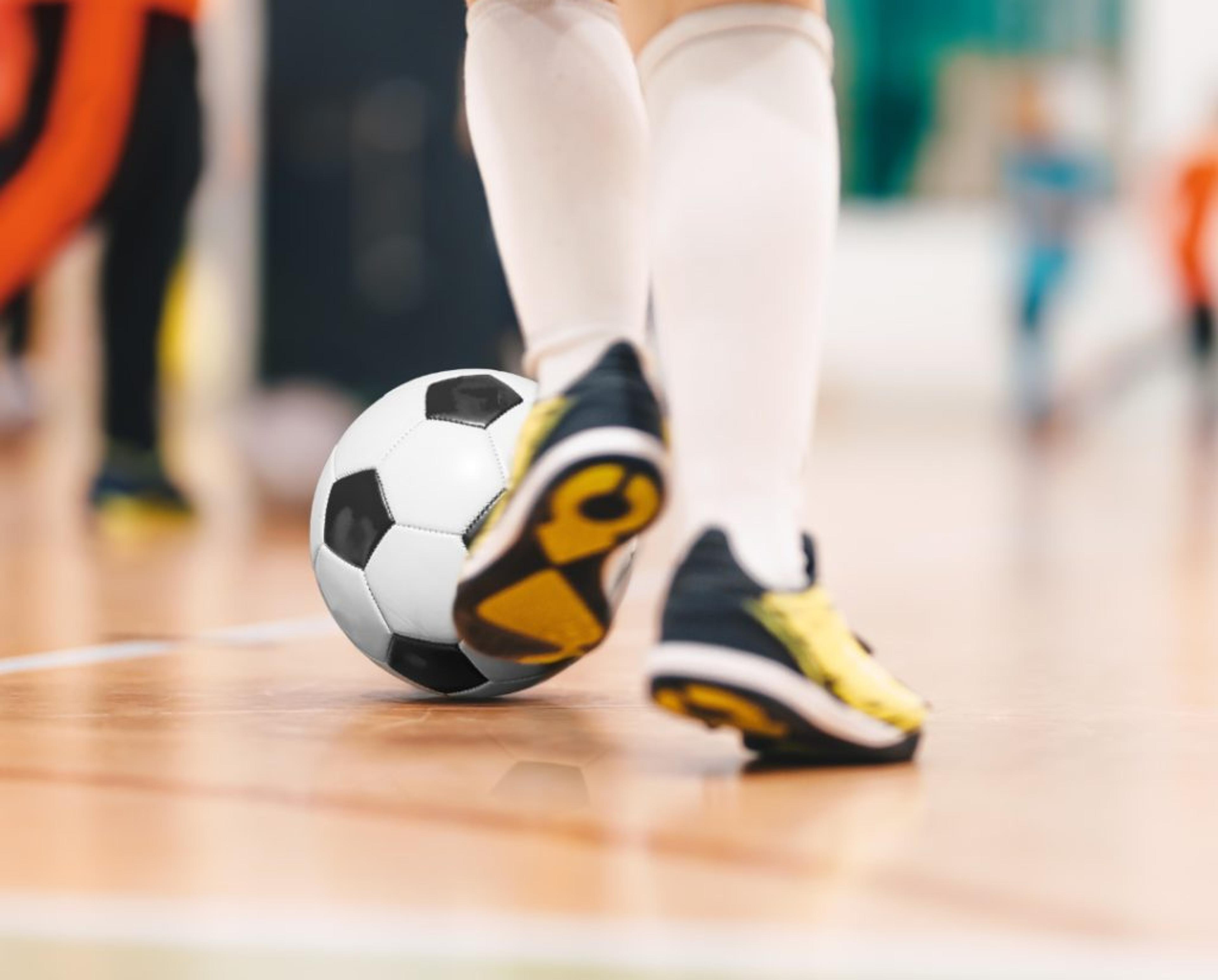 En spelare med vita knästrumpor och svartgula skor står på en inomhusplan med en svartvit fotboll vid fötterna. Andra spelare syns suddigt i bakgrunden.