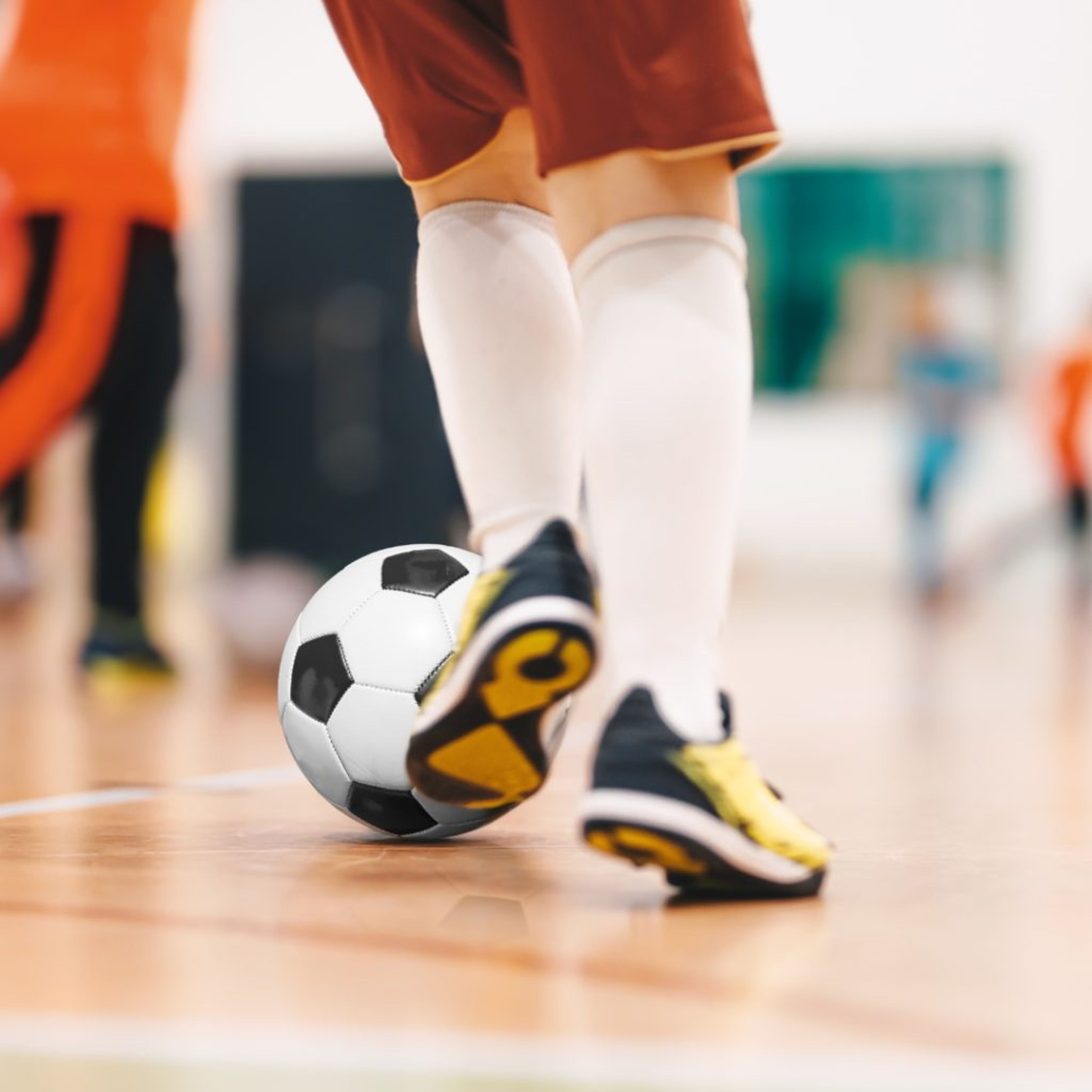 En spelare med vita knästrumpor och svartgula skor står på en inomhusplan med en svartvit fotboll vid fötterna. Andra spelare syns suddigt i bakgrunden.