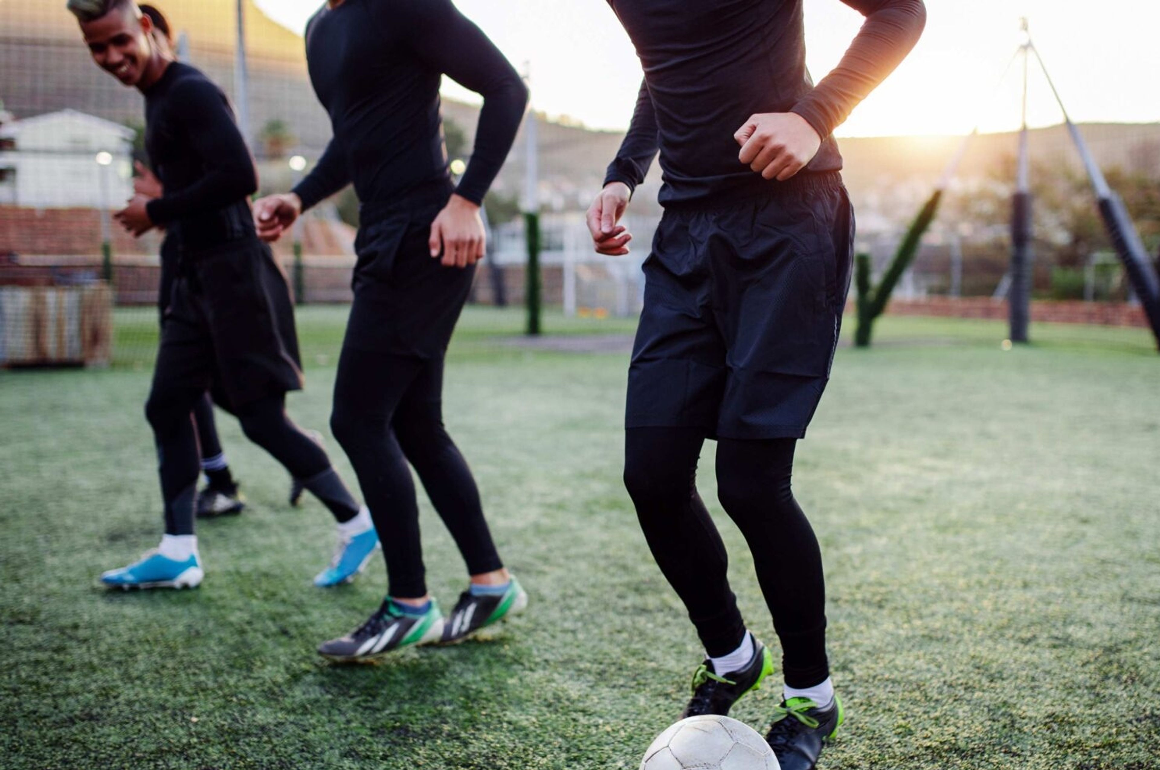 Tre personer tränar fotboll på en grön plan i kvällsljus, alla klädda i svarta träningskläder och sportskor. En vit fotboll syns i förgrunden framför en av spelarna.