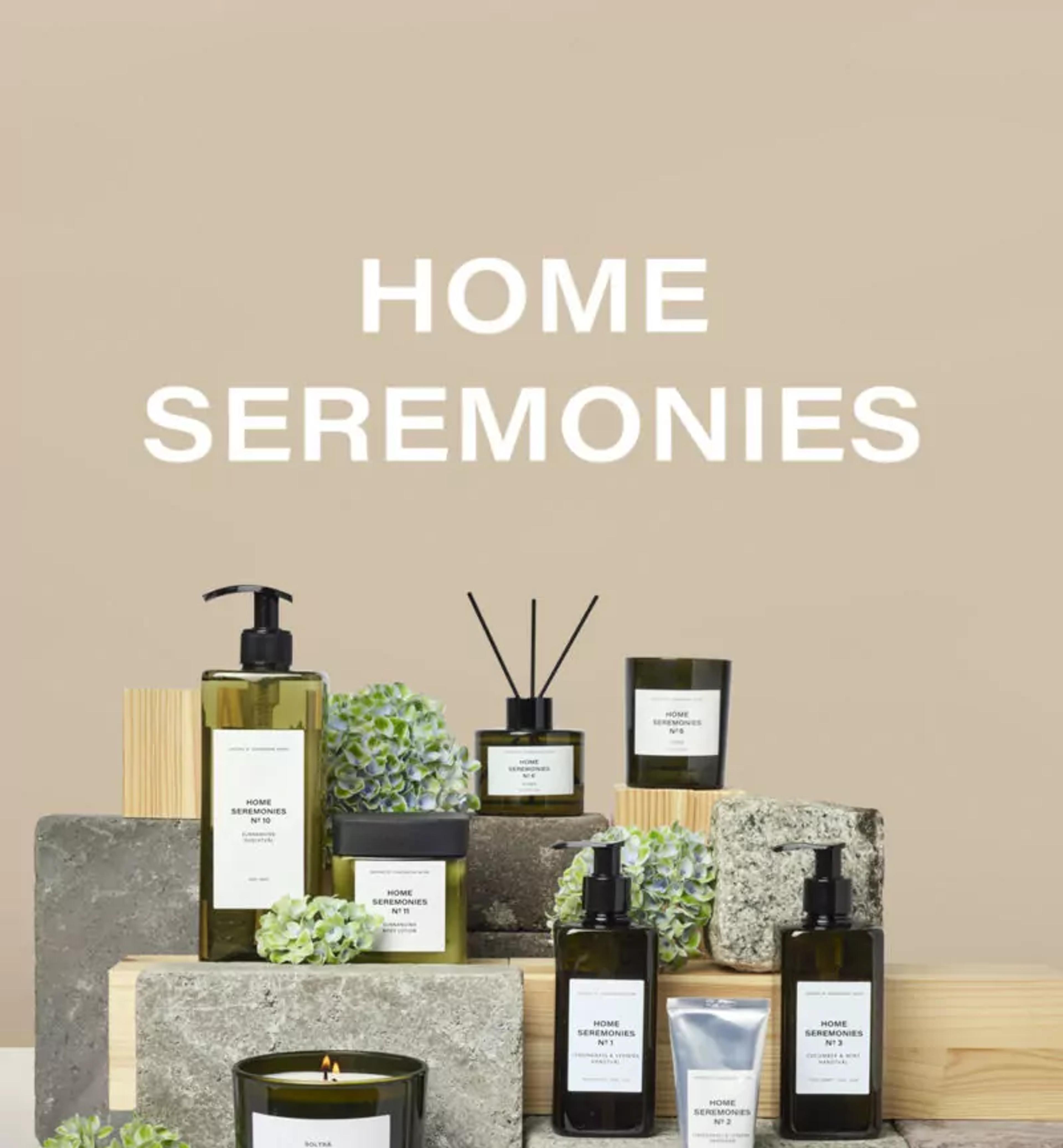 Ett urval av doftljus, handkräm, tvålpump och doftpinnar i stilrena, gröna och svarta förpackningar, arrangerade med stenblock och dekorativa blad. Texten "HOME SEREMONIES" syns tydligt ovanför produkterna