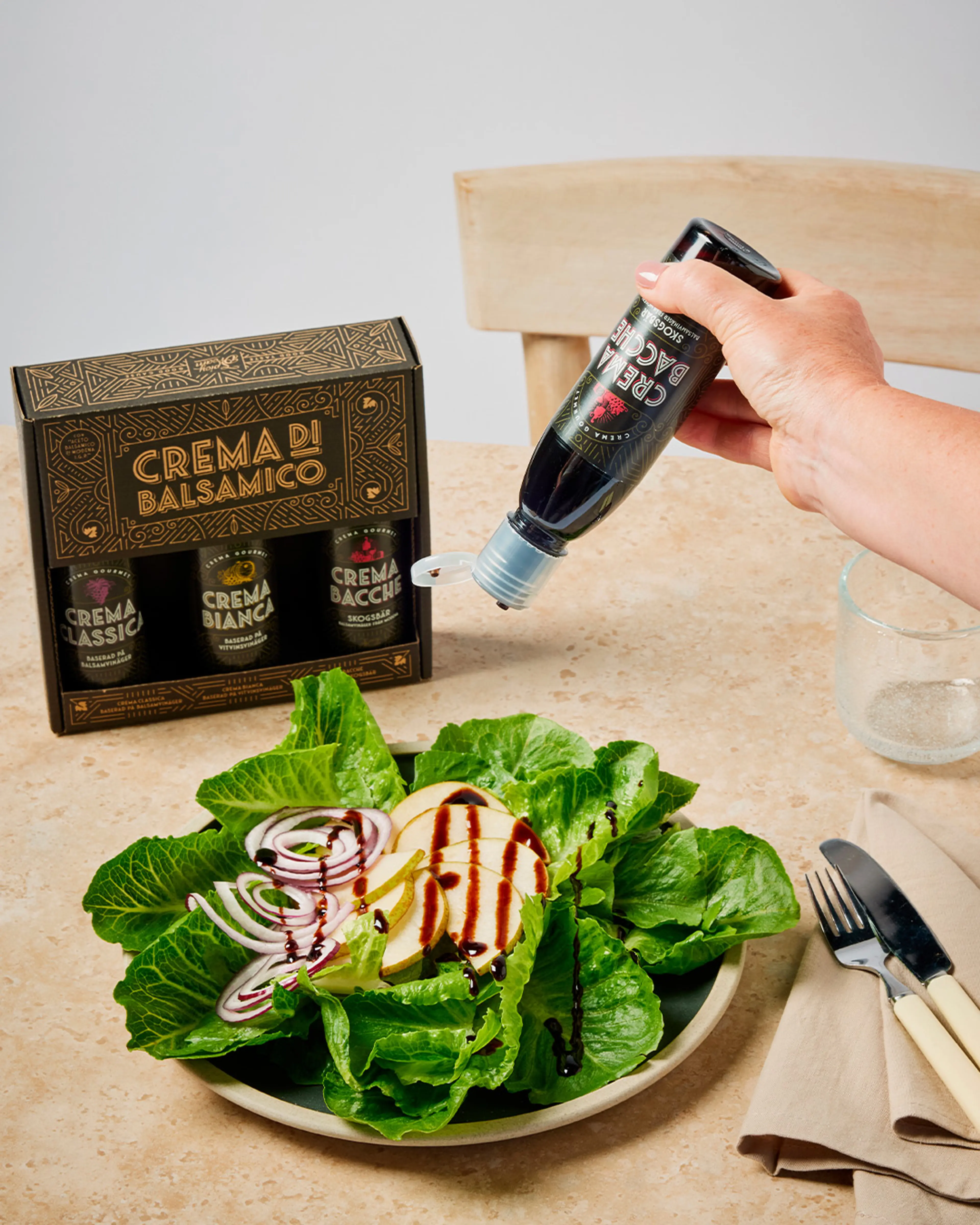En hand häller en mörk balsamicokräm över en fräsch sallad med isbergssallad, rödlök och skivad päron. Tre flaskor balsamicokräm i presentförpackning står i bakgrunden på bordet.