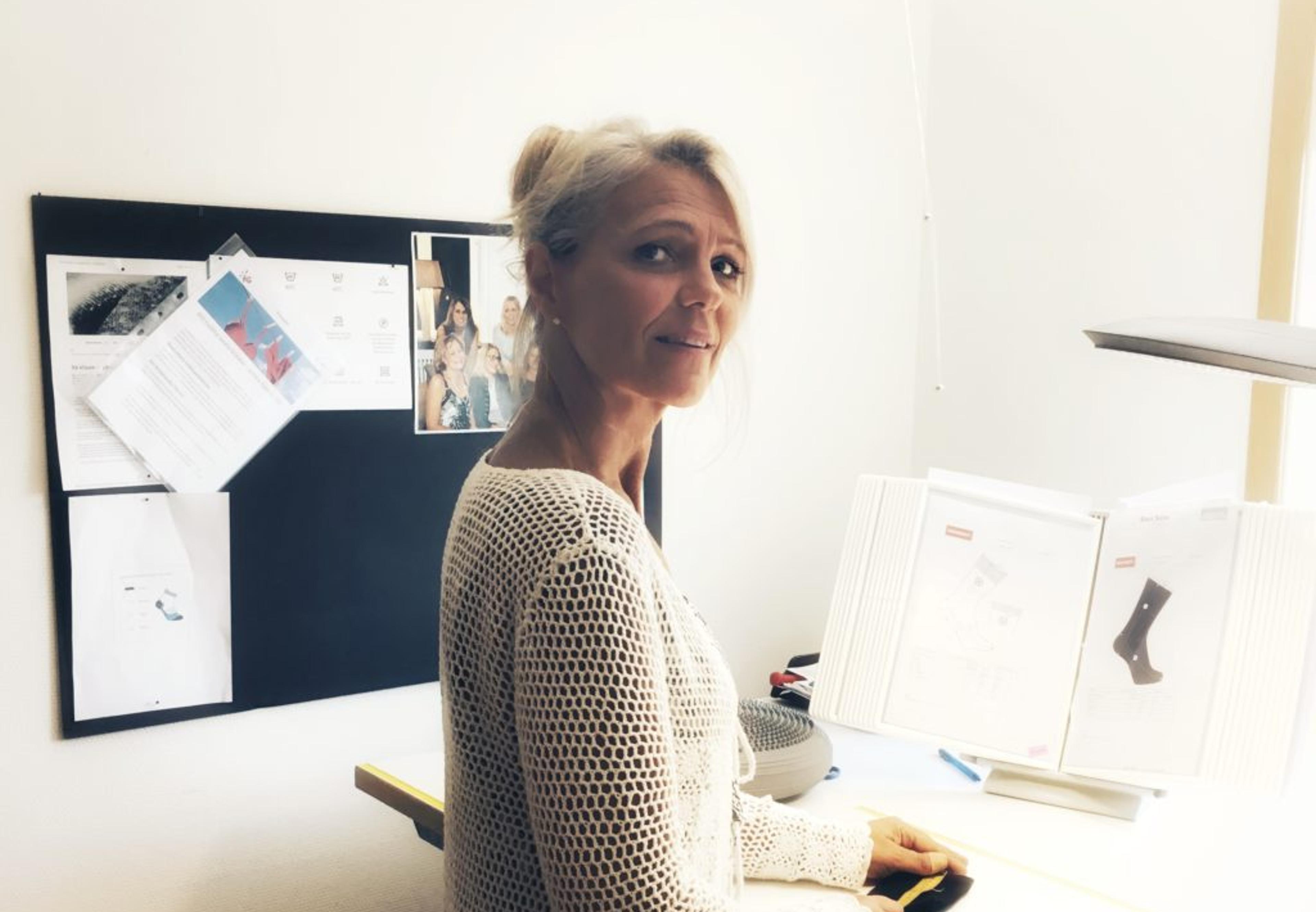 En kvinna i vit, hålmönstrad tröja arbetar vid ett upplyst skrivbord med dokument och ett par svarta strumpor. Bakom henne syns anslagstavla med papper och bilder.