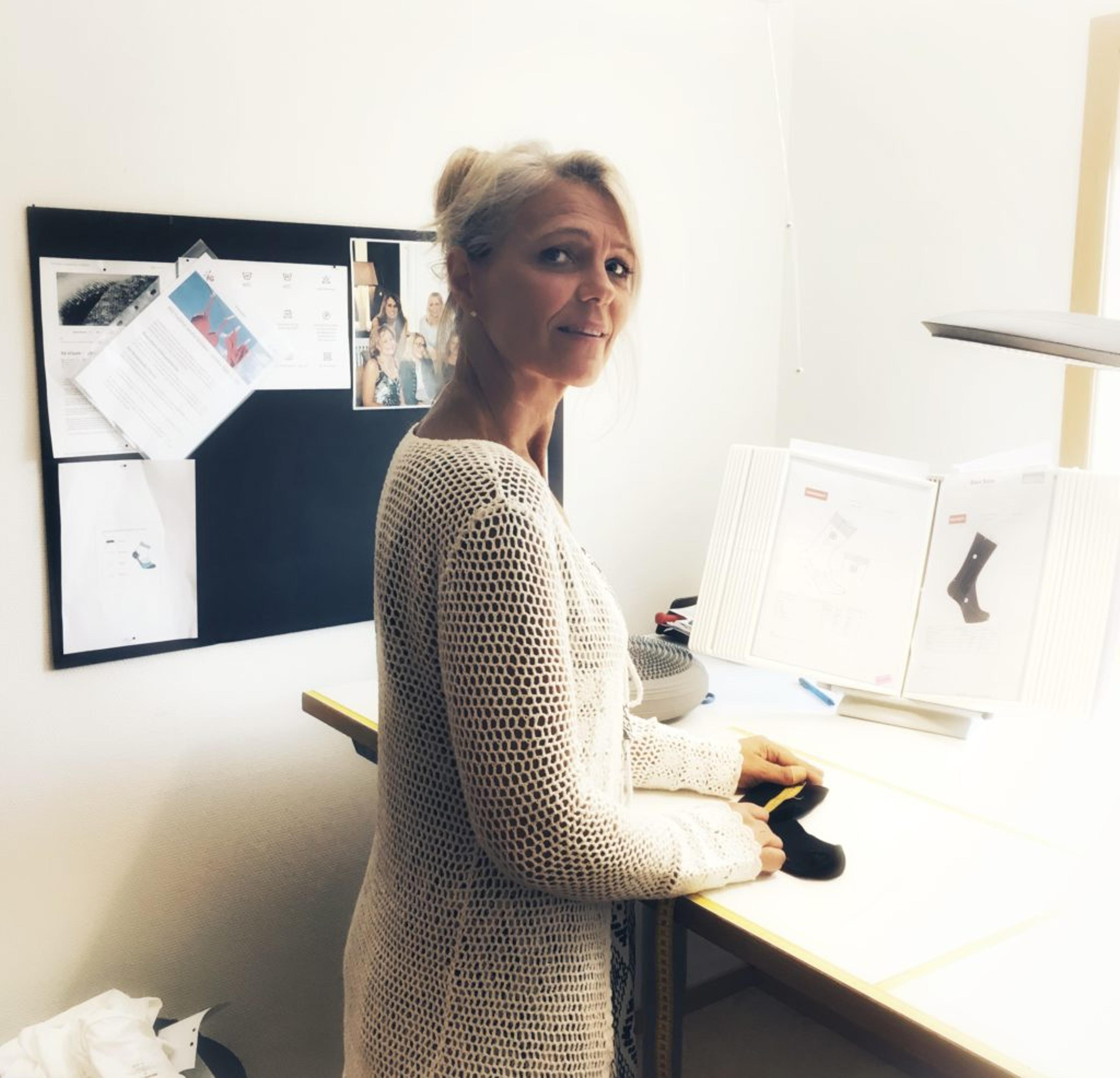En kvinna i vit, hålmönstrad tröja arbetar vid ett upplyst skrivbord med dokument och ett par svarta strumpor. Bakom henne syns anslagstavla med papper och bilder.