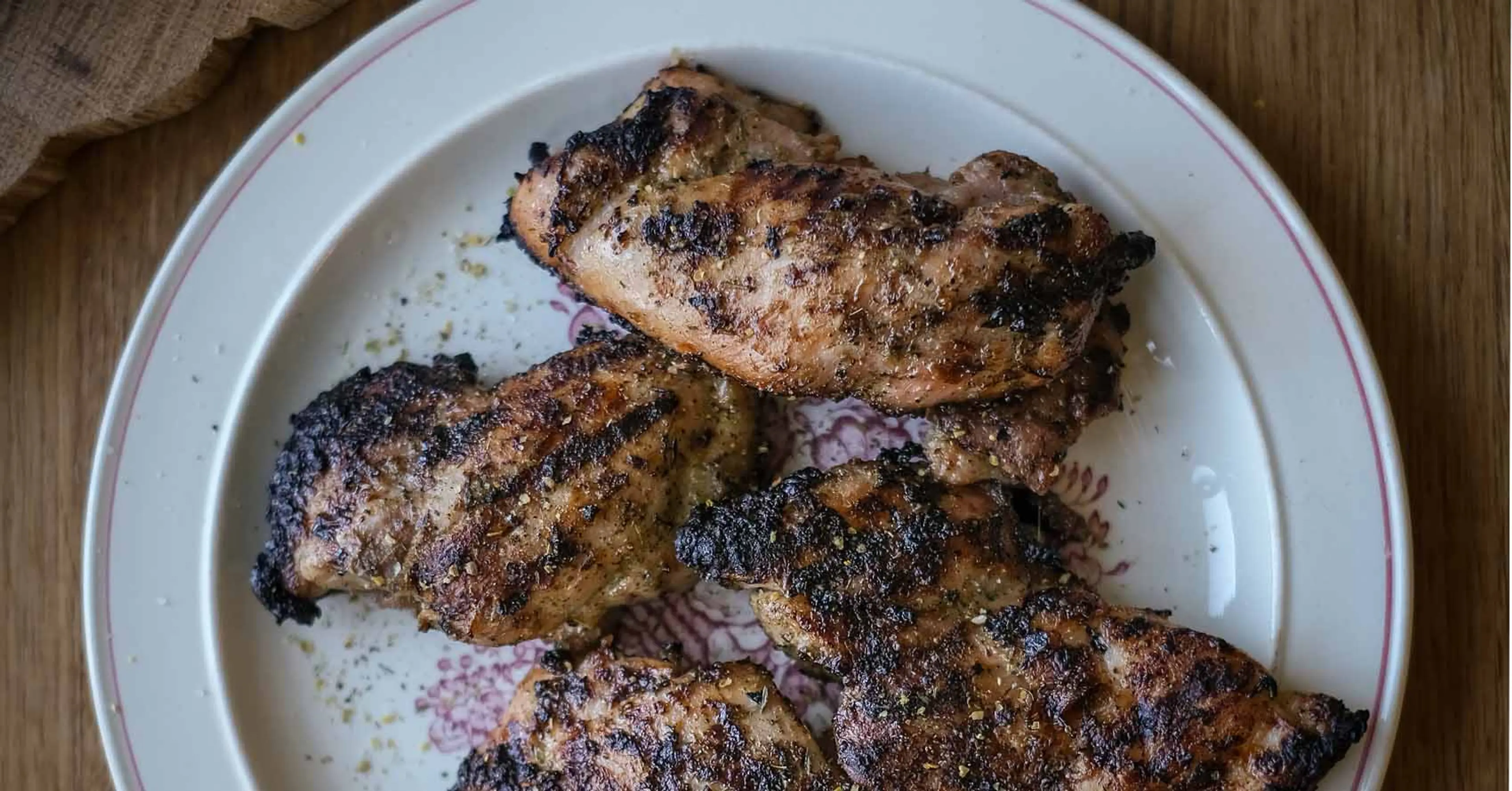 Fem grillade kycklinglårfiléer med en kryddig yta, serverade på en vit tallrik med rosa kant. Ytan är lätt kolgrillad med synliga grillränder.