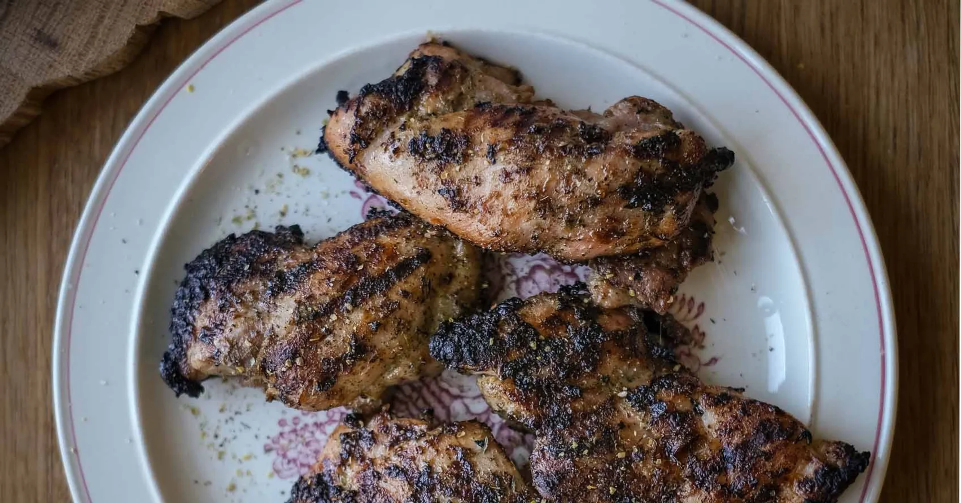 Fem grillade kycklinglårfiléer med en kryddig yta, serverade på en vit tallrik med rosa kant. Ytan är lätt kolgrillad med synliga grillränder.