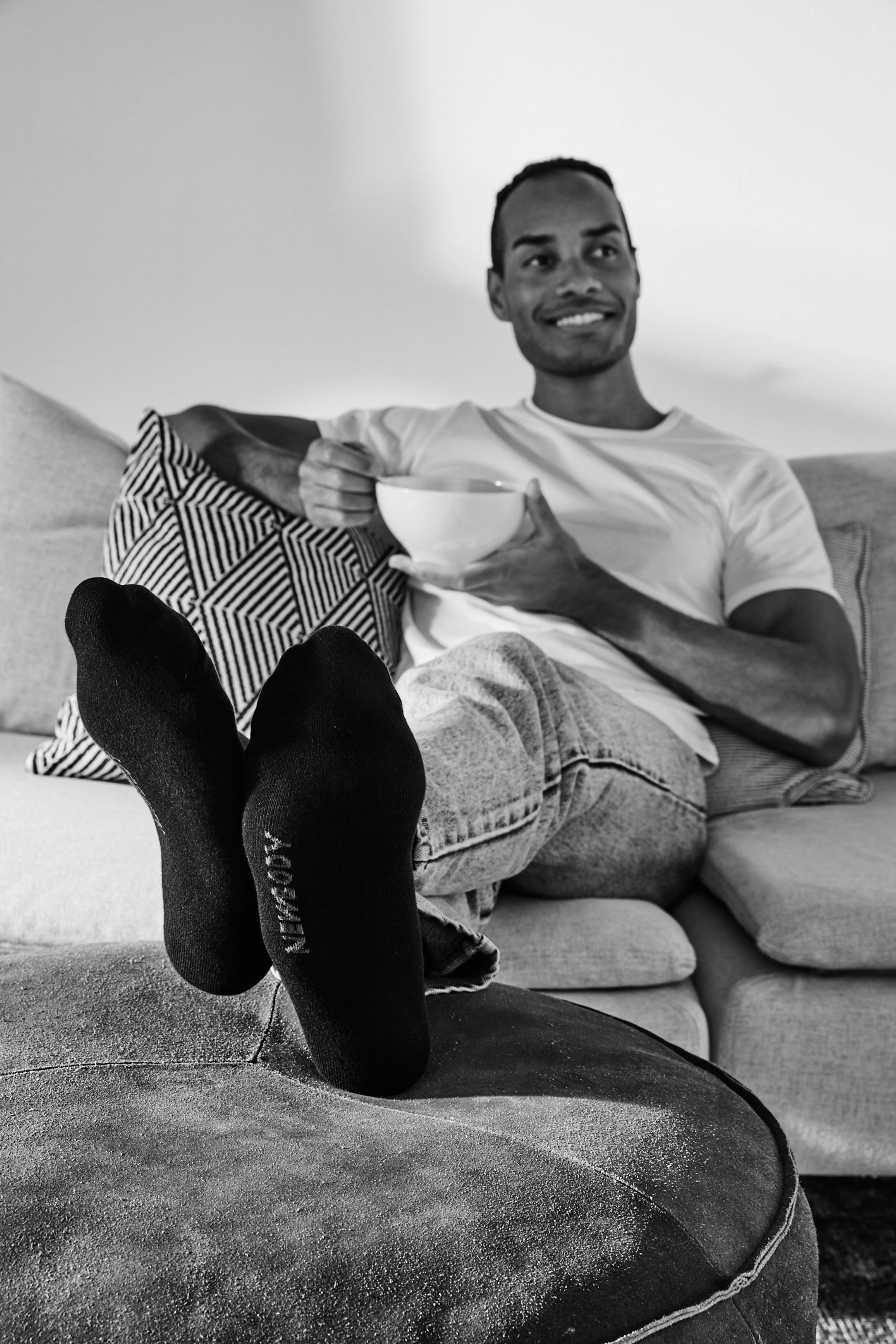 En person sitter avslappnat i en soffa med fötterna vilande på en puff och håller i en stor kopp. Svarta, enkla strumpor omsluter fötterna, med texttryck på undersidan.