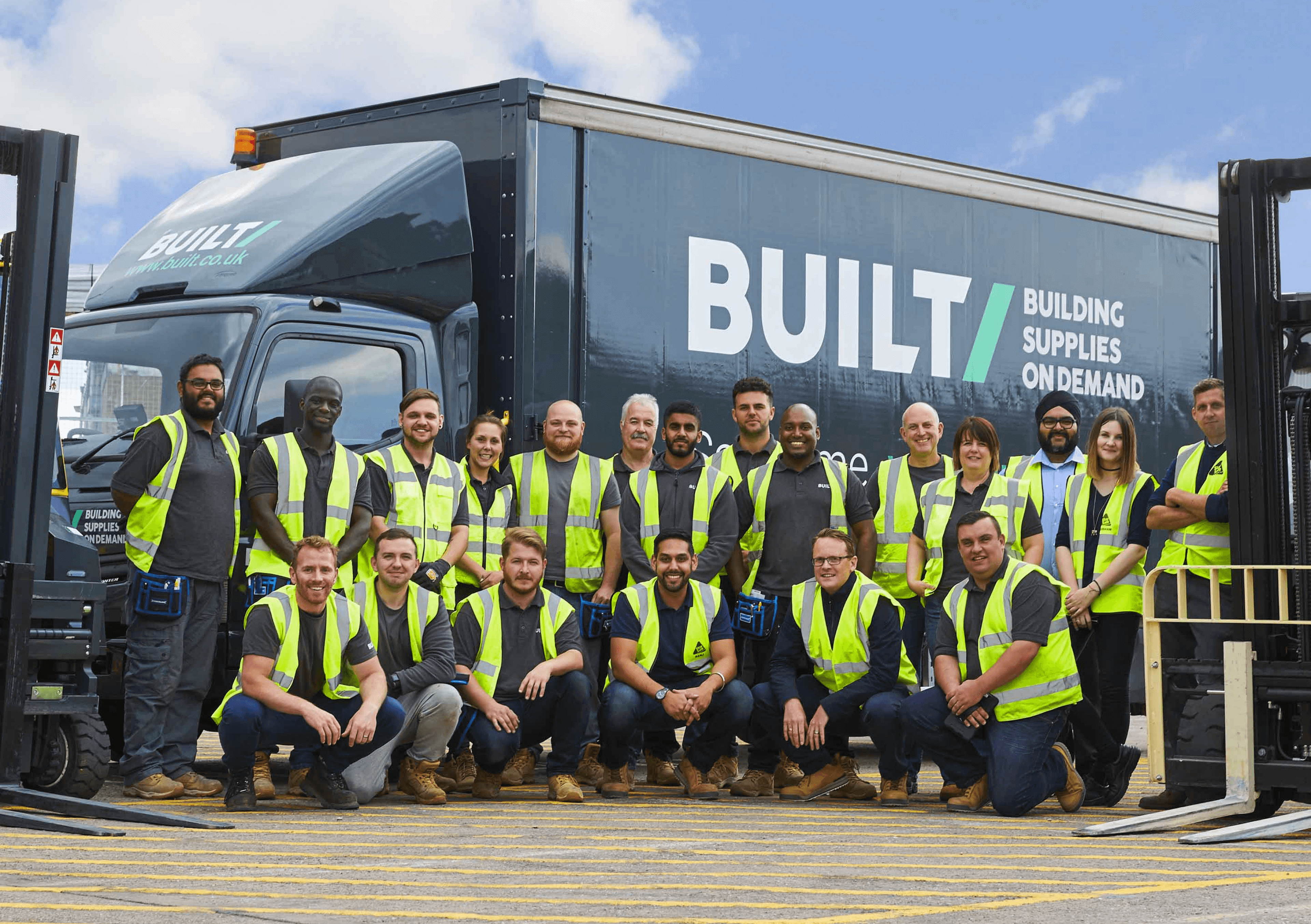 A Built builders merchant team of 20 employees group-shot in-front of a branded truck in an outdoor warehouse