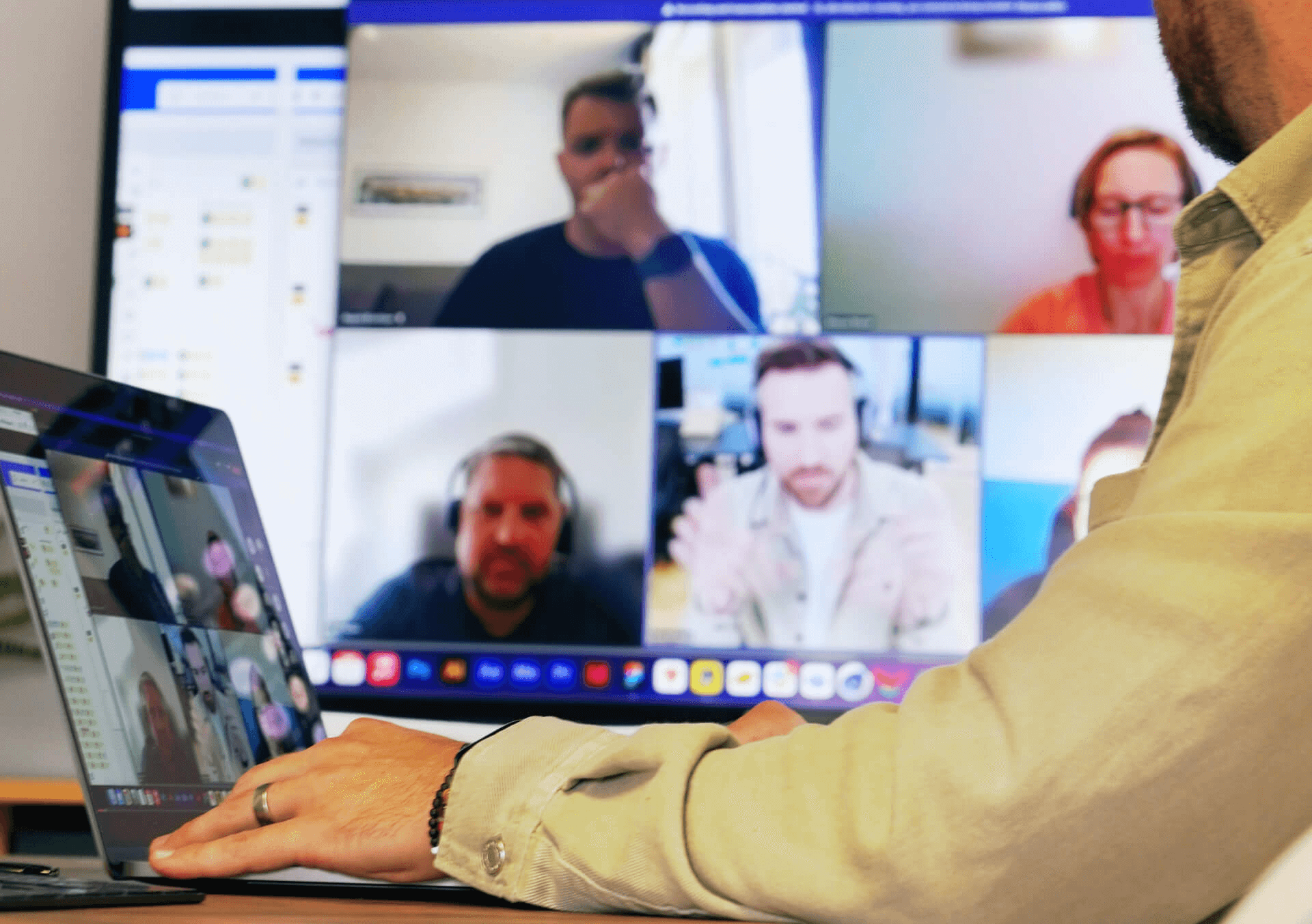 In the foreground a man is on his laptop. The screen behind him shows 5 participants on a product call.