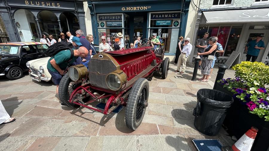 A vintage red FIAT racing car with a brass radiator, exposed chain drive and spoked wheels, drawing a crowd outside G. Horton's pharmacy.