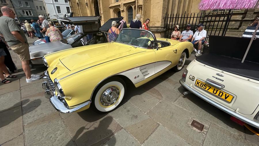 A pale yellow 1958 Chevrolet Corvette convertible on whitewall tyres, parked on the flagstones beside the DeLorean with the church behind.