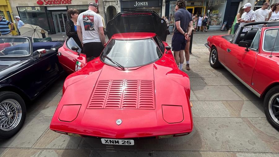 A low, wedge-shaped red Lancia Stratos rally car parked on the Market Place flagstones, with onlookers and shopfronts behind.