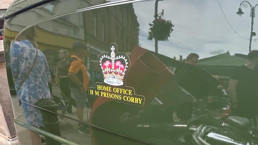A close-up of the van's green flank showing a crown crest reading "Home Office, HM Prisons Corby."