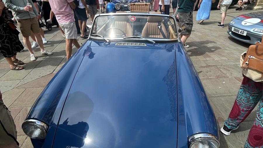 A deep blue MGB roadster with chrome bumpers and a wicker picnic basket strapped to the boot, surrounded by visitors in the Market Place.