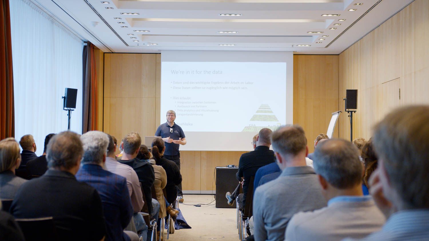 Referent hält einen Fachvortrag vor sitzendem Publikum mit Präsentation an der Leinwand im Eventfilm