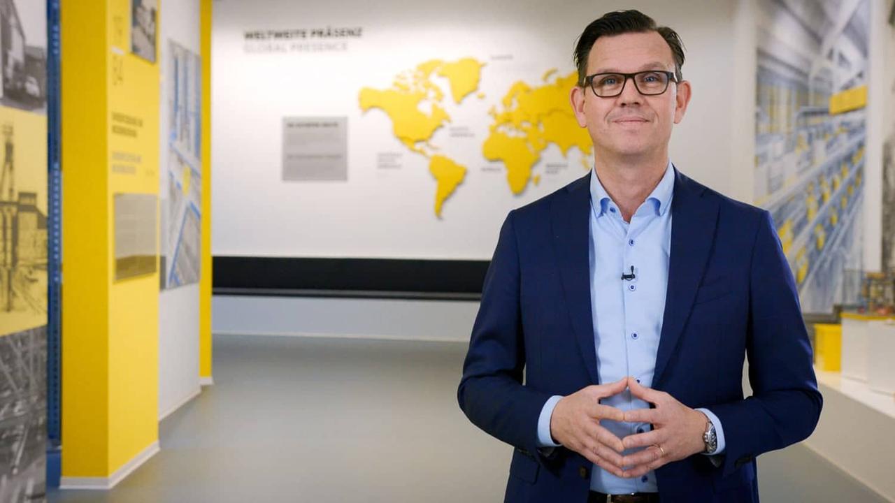 Still aus dem SSI-Schäfer-CEO-Statement: Geschäftsführer im Anzug vor einer Weltkarte im Showroom