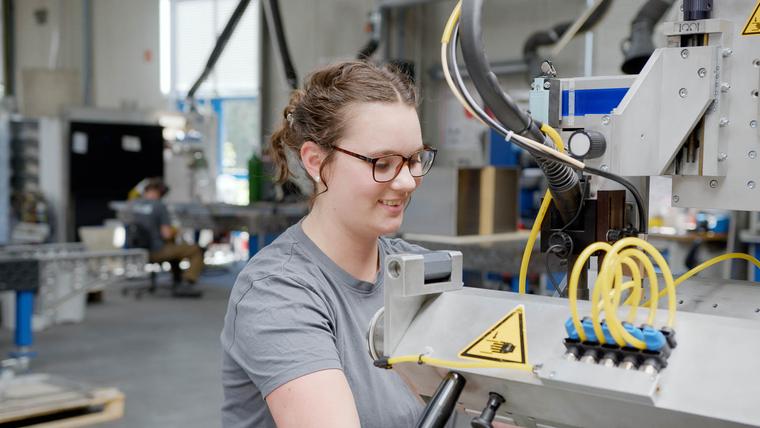 Lächelnde Mitarbeiterin mit Brille bedient eine Fertigungsmaschine in einer Produktionshalle