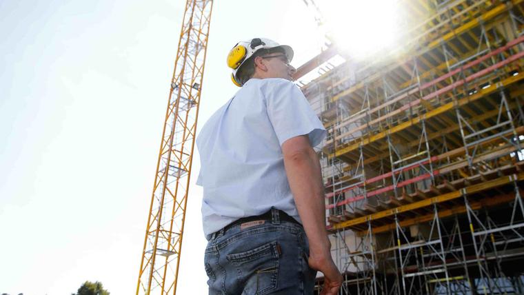 Bauarbeiter mit Helm blickt im Gegenlicht auf Baukran und Gerüst einer Großbaustelle