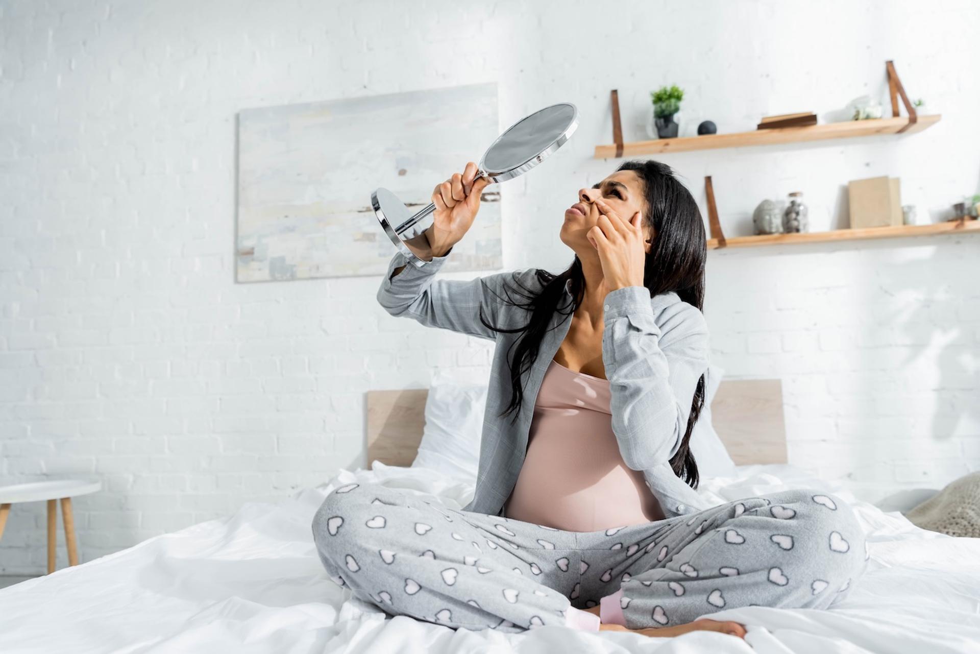 A pregnant woman examines her face in the mirror while sitting in bed